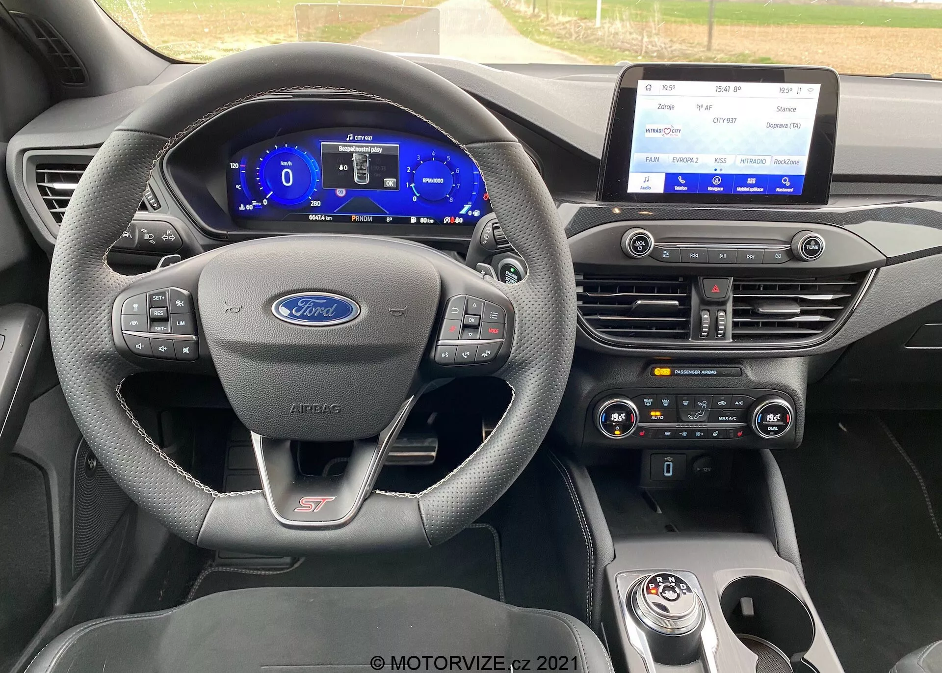 Vista interior de un Ford Focus ST 2022. La imagen se centra en el tablero desde la perspectiva del conductor. El volante es de tres radios con controles multifunción, forrado en cuero con costuras de contraste. Un emblema ST es visible, indicando que es un modelo de alto rendimiento. Detrás del volante, un cuadro de instrumentos digital muestra un velocímetro y un tacómetro con tema azul. La consola central presenta un gran sistema de infoentretenimiento con pantalla táctil horizontal, controles de clima de doble zona y un selector de marchas giratorio con otro emblema ST.