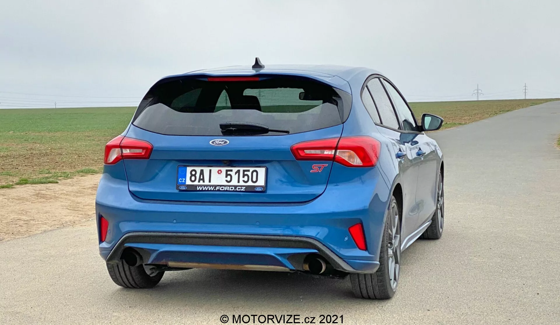 Un hachback azul Ford Focus 2022 se muestra desde un ángulo de tres cuartos trasero en una carretera pavimentada con un cielo nublado de fondo. El auto cuenta con un alerón trasero, luces traseras modernas, una insignia distintiva en la tapa del maletero con una designación alfanumérica debajo, sistema de escape dual, sensores integrados en el parachoques trasero, un difusor trasero, ruedas deportivas de múltiples radios, faldones laterales y un limpiaparabrisas trasero. La imagen destaca el diseño y las características de rendimiento del coche en un posible entorno rural.