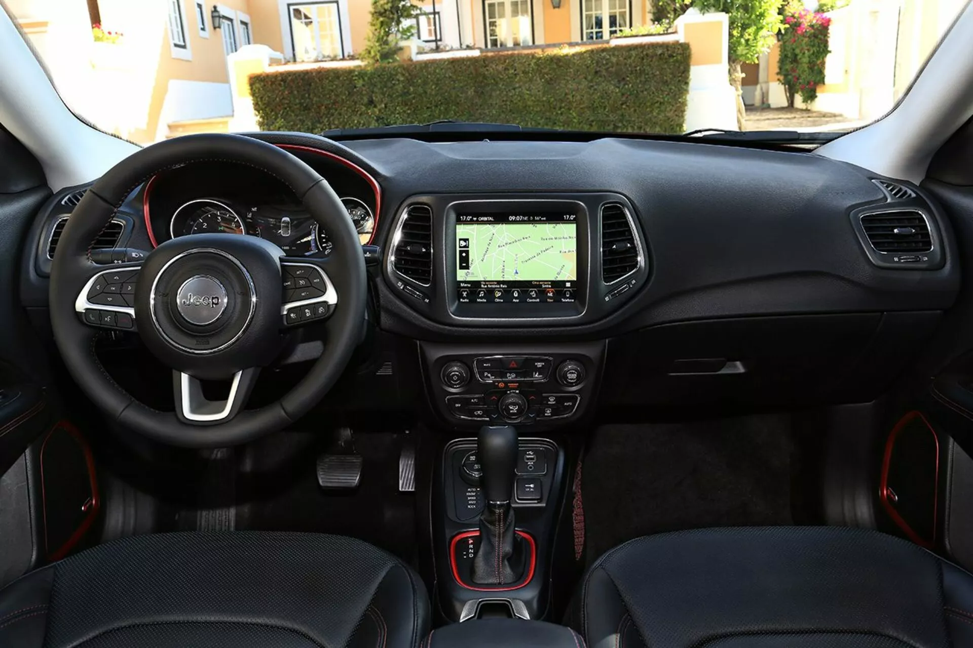 Vista interior del Jeep Compass 2017 desde la perspectiva del conductor mostrando el volante con controles montados, el cuadro de instrumentos, la consola central con sistema de navegación, controles de climatización, palanca de cambios automática con detalles rojos deportivos, botón de encendido-apagado sin llave, y puerto de conectividad multimedia