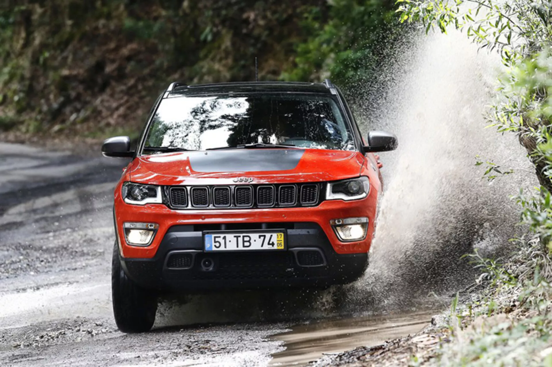 SUV Jeep Compass 2017 en acción dinámica fuera de carretera, mostrado desde un ángulo frontal de tres cuartos enfatizando su robusto diseño y capacidades de rendimiento en un camino forestal embarrado.