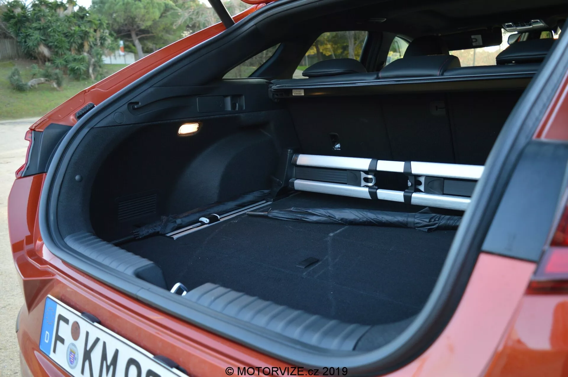 La imagen captura una vista trasera de ángulo de un Kia Ceed 2021 en rojo, centrada en el maletero hatchback abierto. Es el amanecer o el atardecer, y la iluminación suave destaca el espacio de carga vacío y espacioso con un interior negro. Una cubierta de carga está en su lugar, abarcando todo el ancho del maletero. La foto, tomada desde la esquina trasera con un ligero ángulo lateral, también muestra partes del parachoques trasero, las luces traseras y una matrícula parcialmente visible. El maletero tiene un piso de carga plano, una fuente de luz a la izquierda y un compartimento lateral a la derecha.