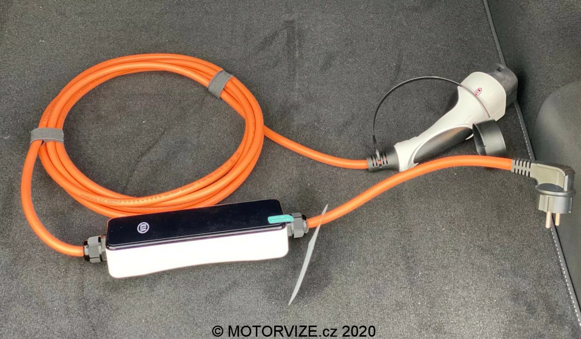 Cable de carga y adaptador para vehículo eléctrico (EV) del Kia Ceed 2018, con un EVSE (Equipo de Suministro de Vehículo Eléctrico) portátil naranja de alta visibilidad para seguridad. La configuración incluye un conector y enchufe de carga para el uso en tomacorrientes domésticos, así como una caja de control en línea con indicadores LED, todo colocado en la superficie alfombrada del maletero o área de carga del vehículo.