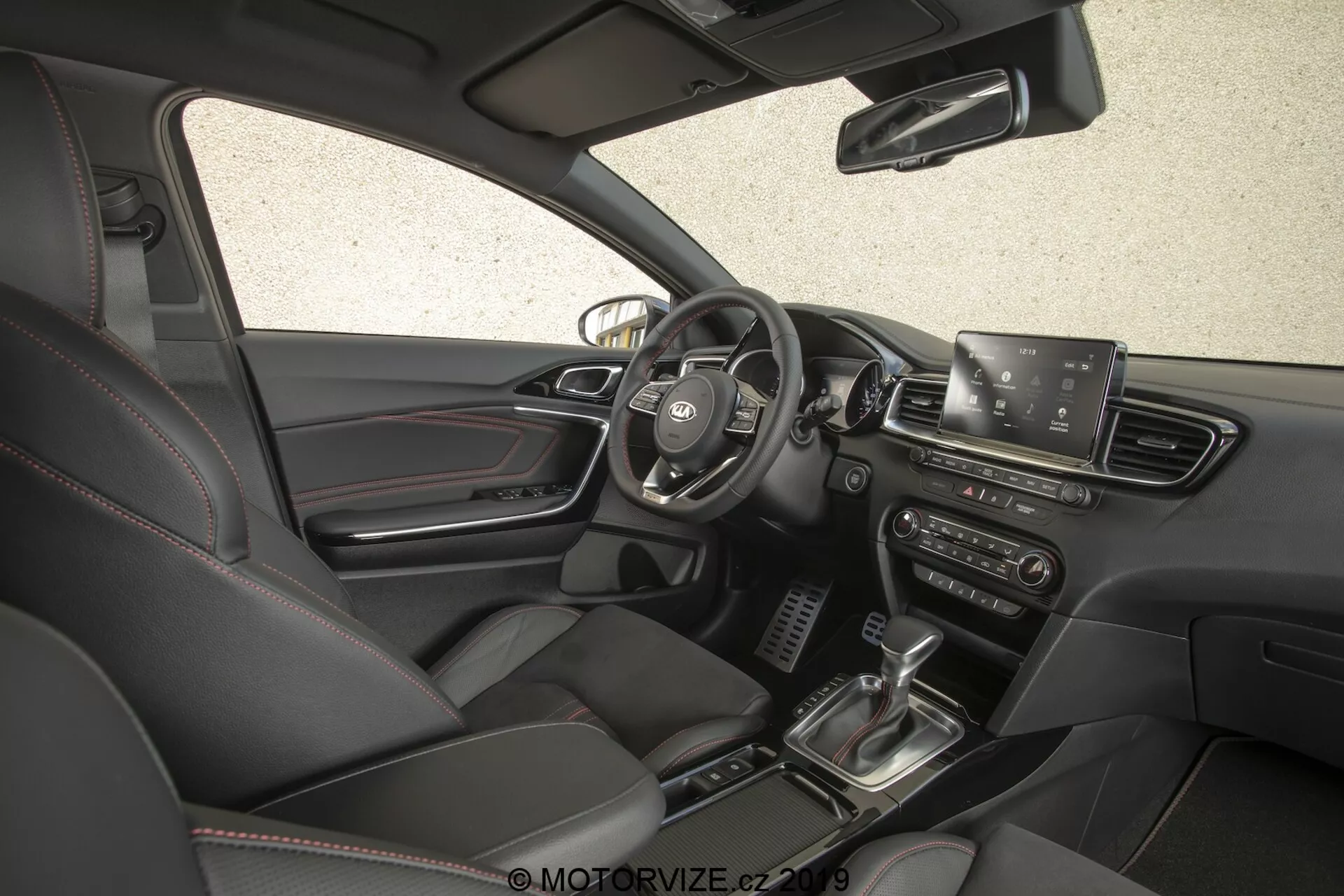 Vista interior del coche Kia Ceed 2021, mostrando el lado del conductor desde un ángulo alto y ligeramente hacia adelante. La imagen, capturada desde el lado del pasajero, proporciona una vista completa del tablero, incluyendo el volante central con botones de control, visibilidad parcial del panel de instrumentos, una pantalla de infoentretenimiento destacada, rejillas de ventilación, botones y perillas de control de clima, palanca de cambios, sistema de arranque con botón, asientos de cuero con controles de ajuste eléctrico y una paleta de colores interiores oscuros.