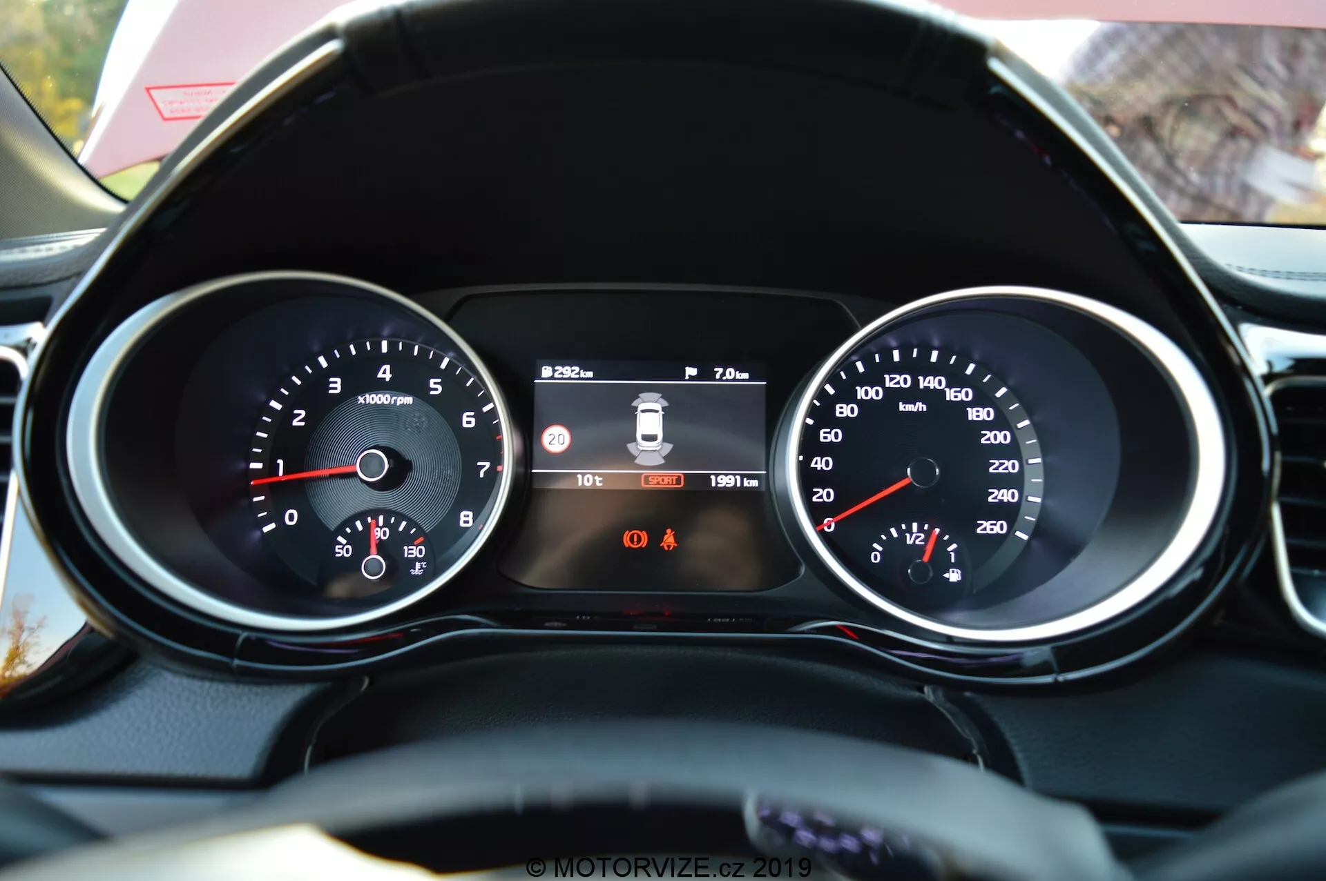 Vista en primer plano desde la perspectiva del conductor del panel de instrumentos del Kia Ceed 2021 y parte del volante terminado en cuero. Las características incluyen un tacómetro a la izquierda con lecturas de RPM, un display digital central que muestra el sistema de advertencia de salida de carril, la hora y el odómetro, y un velocímetro a la derecha con la velocidad en km/h. Debajo del display central, se ven dos luces indicadoras de advertencia, siendo identificable el recordatorio del cinturón de seguridad.