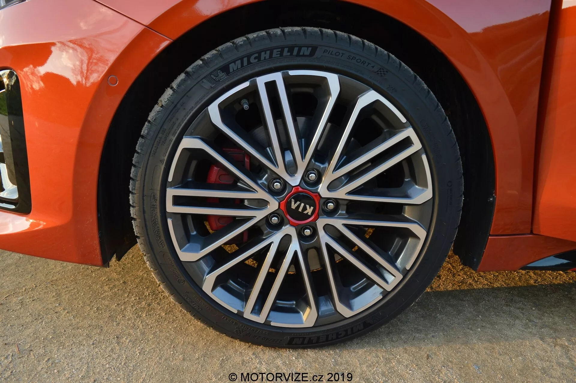 Primer plano de la rueda delantera del lado del pasajero de un Kia Ceed 2021, en un ángulo diagonal leve, mostrando una llanta de aleación multirradio con secciones internas más oscuras y bordes de máquina brillantes. Presenta un buje con marca y una marca roja 'VT1', neumático Michelin con información detallada en la pared lateral y una pinza de freno roja que indica componentes orientados al rendimiento. Las sombras suaves y los reflejos sugieren que la foto fue tomada con luz natural, probablemente durante la hora dorada.