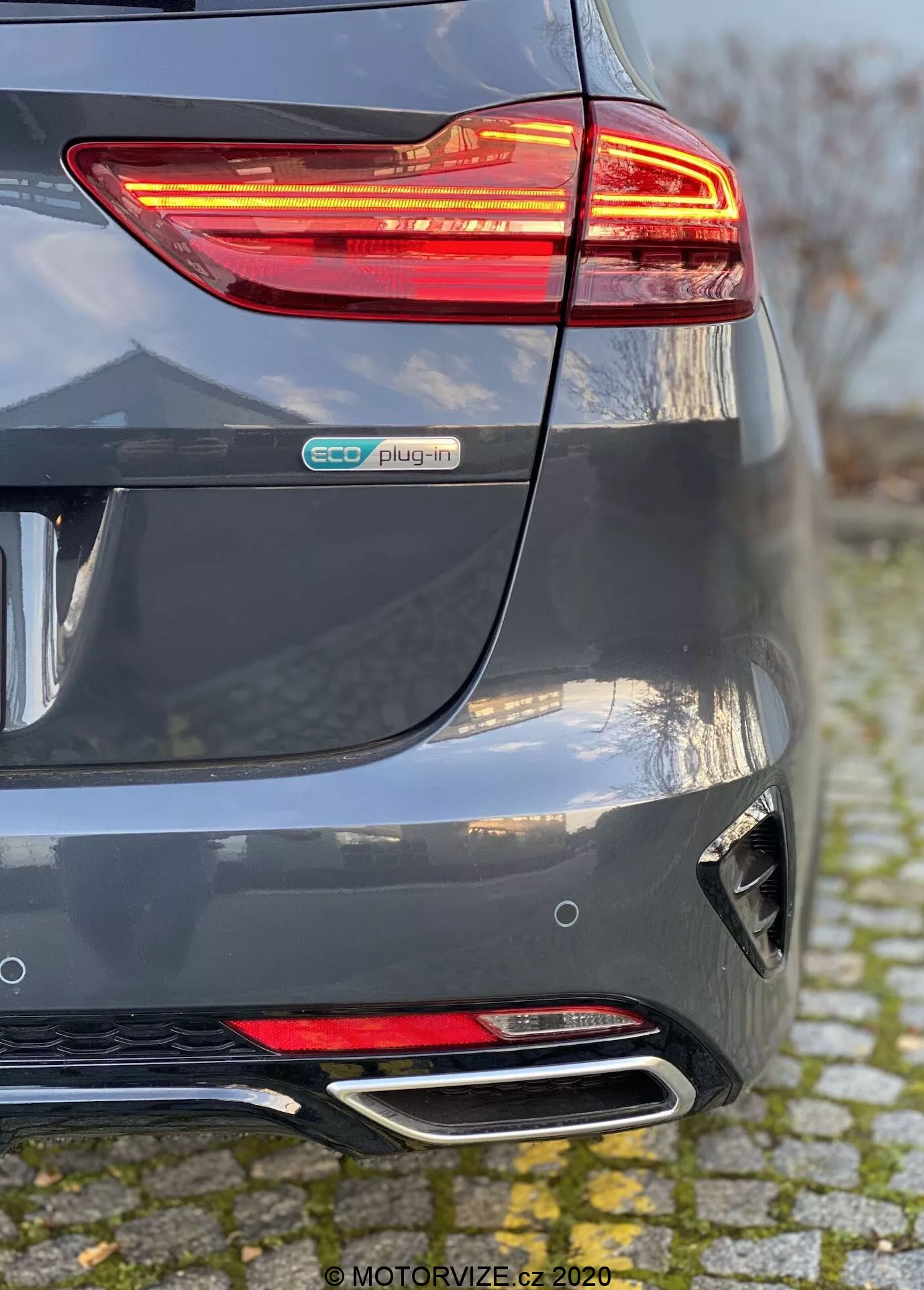 Vista detallada de la sección trasera de un Kia Ceed 2018 en gris metálico, destacando la luz trasera LED iluminada, la insignia eco plug-in, el paragolpes trasero esculpido con reflector y difusor, y los tubos de escape cromados dobles. La perspectiva muestra la convergencia del parachoques trasero y el lado derecho, enfatizando los elementos del vehículo eléctrico híbrido y las características de diseño moderno.