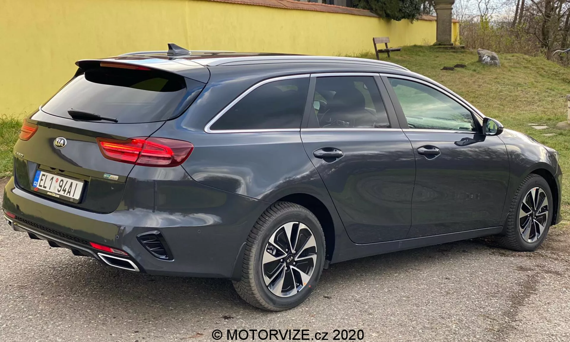 Un Kia Ceed 2018 metálico gris oscuro, mostrado desde una vista trasera de tres cuartos, destacando su diseño moderno de hatchback o familiar con ventana trasera inclinada, alerón trasero integrado, llantas de aleación multirradio, manijas de puertas y espejos laterales del color de la carrocería probablemente con señales de giro integradas, distintivo conjunto de luces traseras horizontales posiblemente con tecnología LED, antena de aleta de tiburón, doble punta de escape, tercera luz de freno montada en alto, capacidad de descongelación de la ventana trasera, sensores o reflectores indicativos de un sistema de asistencia de estacionamiento trasero, fotografiado de cerca con iluminación neutra.