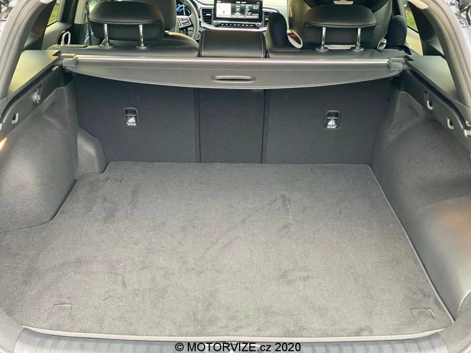 Vista cercana del maletero del Kia Ceed 2018 desde una perspectiva trasera, destacando el área de carga espaciosa y limpia con una superficie alfombrada. La puerta del maletero está levantada, revelando una cubierta retráctil, respaldos de los asientos traseros de color oscuro, probablemente de cuero, y pestillos plateados para abatir los asientos. Los lados están revestidos con un material suave y hay un pequeño compartimento visible en el lado derecho. Imagen tomada desde un ligero ángulo, mostrando el diseño y los materiales del interior del maletero del coche.