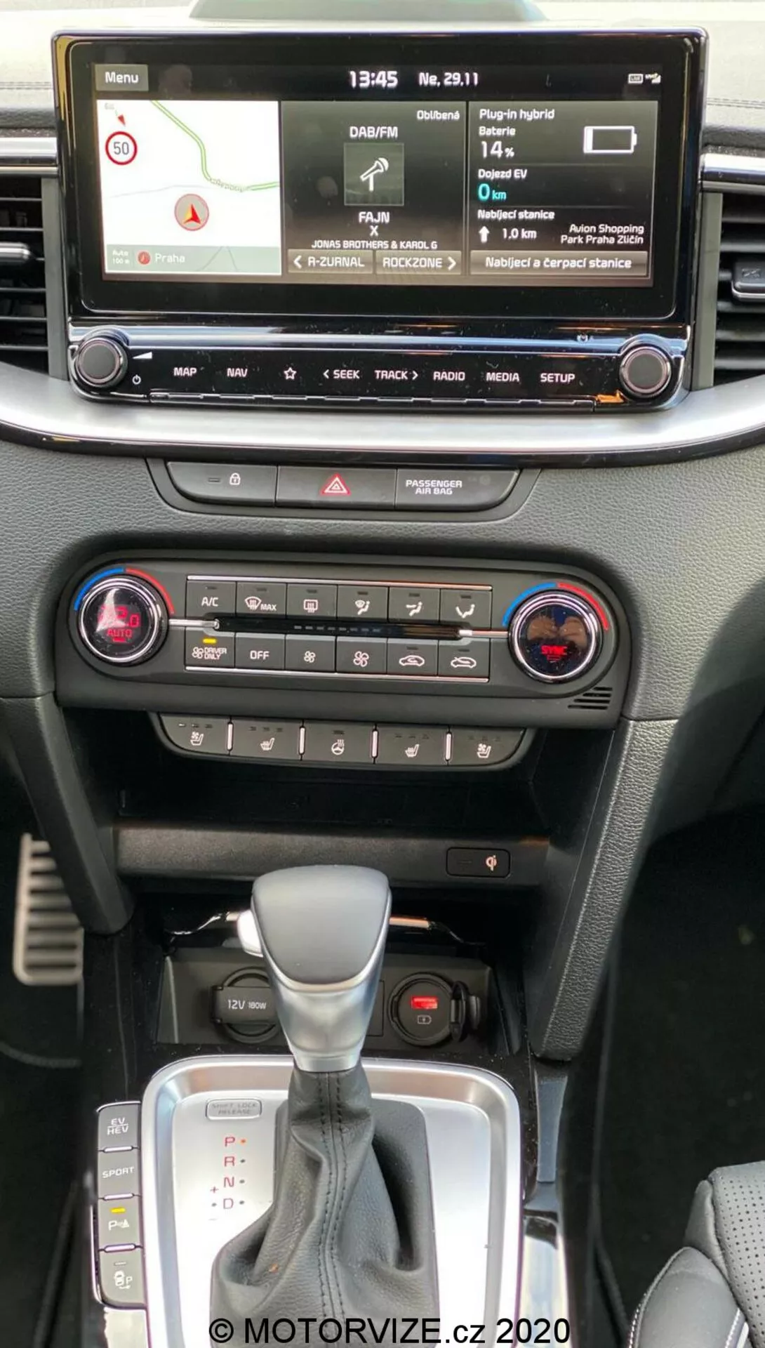 Interior del Kia Ceed 2018 que muestra la consola central desde la perspectiva del pasajero en la fila delantera, con pantalla de infoentretenimiento digital con mapa de navegación, reproductor de medios, configuraciones del vehículo, controles de clima con sistema de doble zona, botón de luces de emergencia, palanca de cambios automática con funda de cuero y ranura para llavero de arranque sin llave.