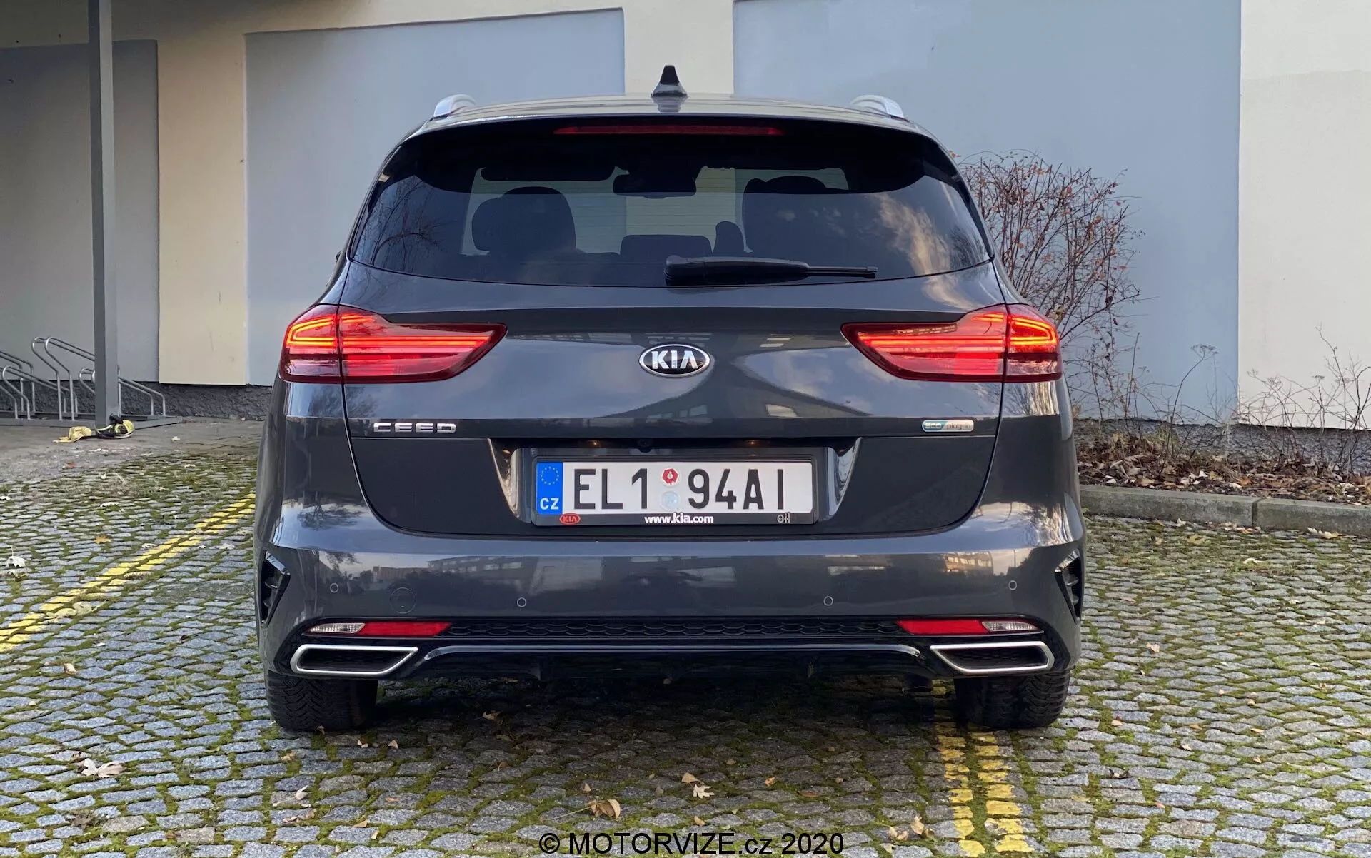 Vista trasera de un Kia Ceed 2018 estacionado en un suelo de adoquines cerca de un bordillo. El coche, capturado durante el día desde un ángulo recto al nivel de los ojos, muestra sus luces traseras LED divididas, parabrisas trasero con limpiaparabrisas, emblema del fabricante, nombre del modelo, placa de matrícula, parachoques trasero con reflectores y sensores de estacionamiento, sistema de doble escape y acabado metálico gris oscuro. El fondo incluye un edificio, indicando una zona residencial o comercial.