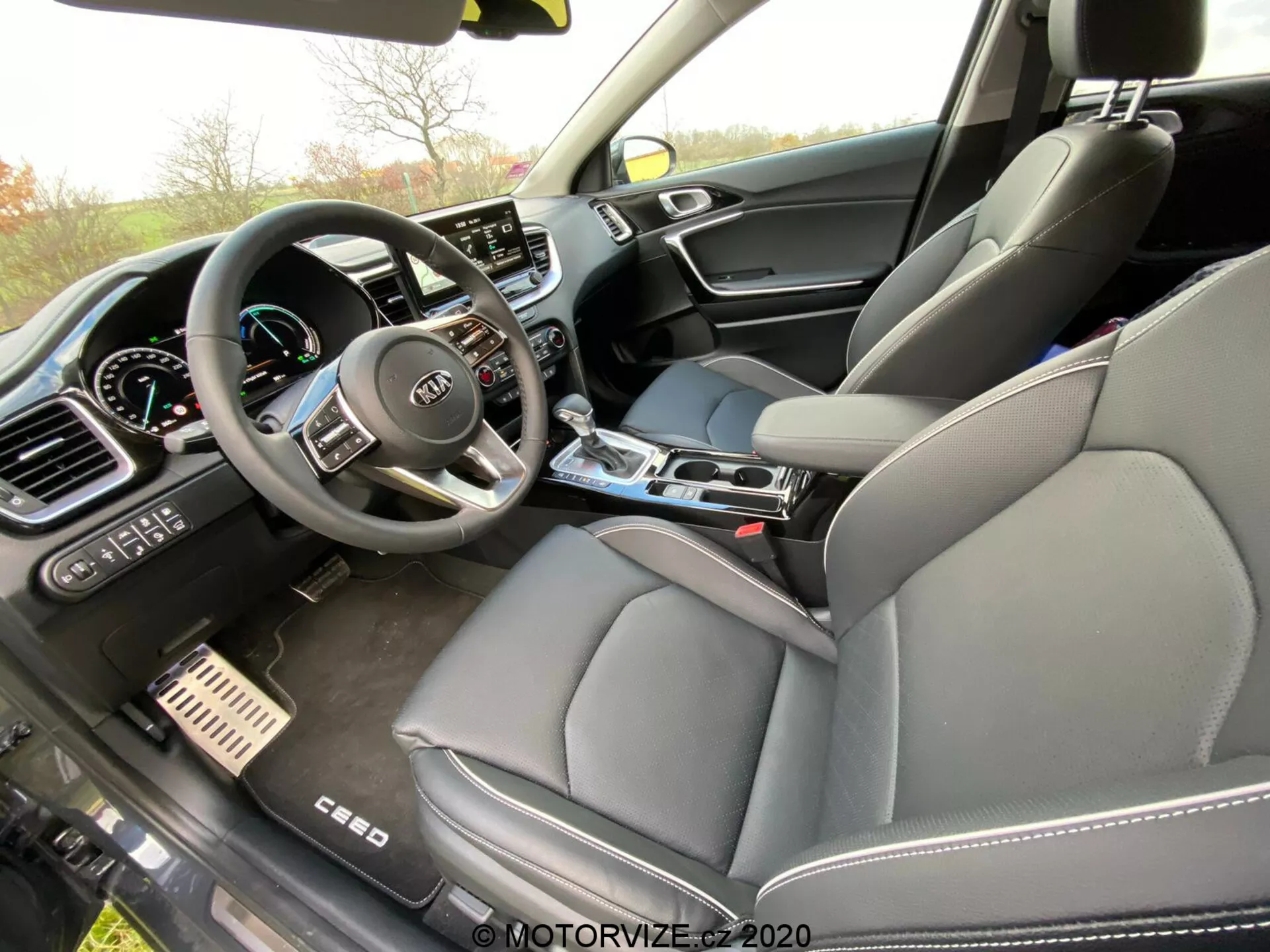 Vista del interior de un Kia Ceed 2018 desde la perspectiva del pasajero, capturando el compartimento delantero con enfoque en el tablero, consola central y asiento del conductor. El volante con múltiples botones de control y el logotipo de Kia es visible, junto con un cuadro de instrumentos digital parcialmente visible. La parte central del tablero cuenta con una gran pantalla táctil de infoentretenimiento rodeada de ajustes manuales de control de clima. La consola central incluye una palanca de cambios, portavasos y diales de control. También se observa el asiento del pasajero delantero con tapicería de cuero y un panel de puerta de materiales mixtos, lo que sugiere un nivel de acabado lujoso. Los pedales de freno y acelerador con superficie metálica indican un toque deportivo premium.