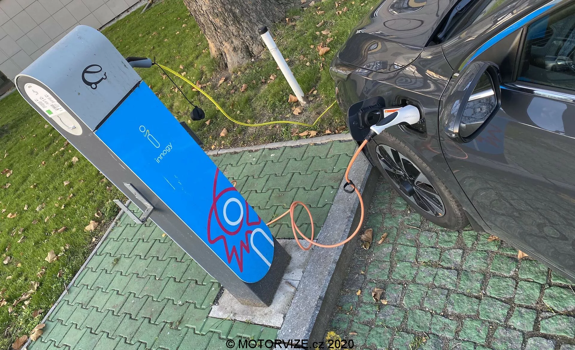 Vista aérea de un vehículo eléctrico moderno cargándose en una estación de carga pública. La imagen captura la sección trasera derecha del coche, centrando en el puerto de carga con el cable naranja grueso conectado. La estación de carga presenta una fachada azul con un gráfico de chispa eléctrica y una pantalla en la parte superior, sugiriendo elementos de interfaz de usuario. La escena está situada en un entorno verde parecido a un parque, indicando una ubicación urbana o suburbana conveniente.
