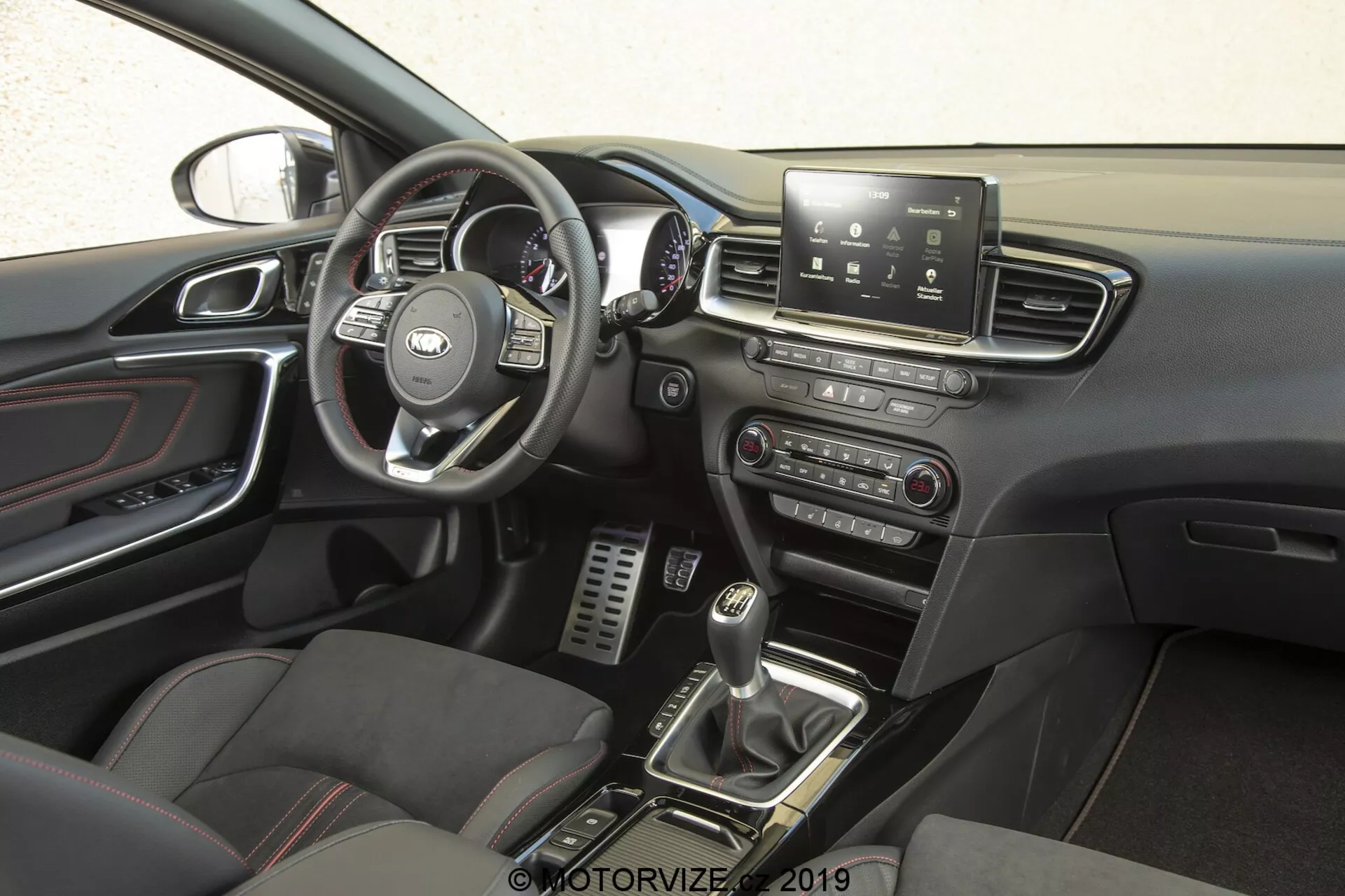 El interior del Kia Ceed 2021, centrado en el lado del conductor desde una perspectiva lateral. El volante, con el logotipo reconocible del fabricante, cuenta con controles multifuncionales para gestionar el sistema de infoentretenimiento, control de crucero, conectividad telefónica y posiblemente comandos de voz. Detrás, se pueden ver el velocímetro, tacómetro y medidores de información del conductor. La consola central tiene una pantalla táctil de infoentretenimiento que ofrece navegación, configuración del vehículo, reproducción de medios y conectividad Bluetooth. Debajo, hay controles manuales para los sistemas de audio y clima. La palanca de cambios de la transmisión manual, rodeada de botones para modos de conducción o control de tracción, y los asientos deportivos con inserciones de cuero y tela, realzan la sensación de rendimiento premium. Los pedales metálicos en el espacio para los pies del conductor sugieren un énfasis en el rendimiento, mientras que los detalles cromados en las salidas de aire, asas de las puertas y otros detalles añaden sofisticación. El panel de la puerta incluye controles para las ventanas eléctricas y posiblemente ajustes de los espejos eléctricos, y una parte del retrovisor es visible, lo que sugiere funciones como atenuación automática o una brújula incorporada.