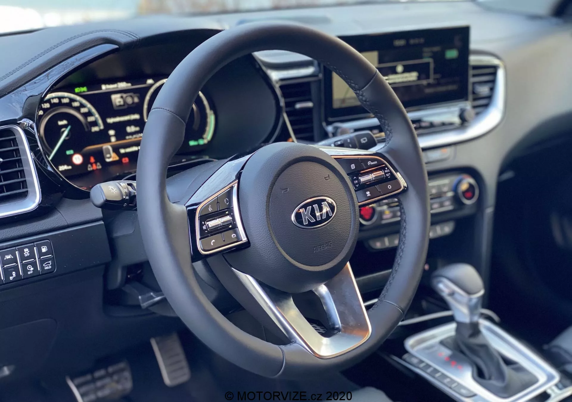 Vista en primera persona del interior del Kia Ceed 2018 destacando el volante multifuncional de tres radios con logotipo de Kia y el cuadro de instrumentos digital, mostrando velocímetro y posible tacómetro, junto con el sistema de infoentretenimiento con pantalla táctil, controles de climatización, palanca de cambios automática plateada y consola central detallada.