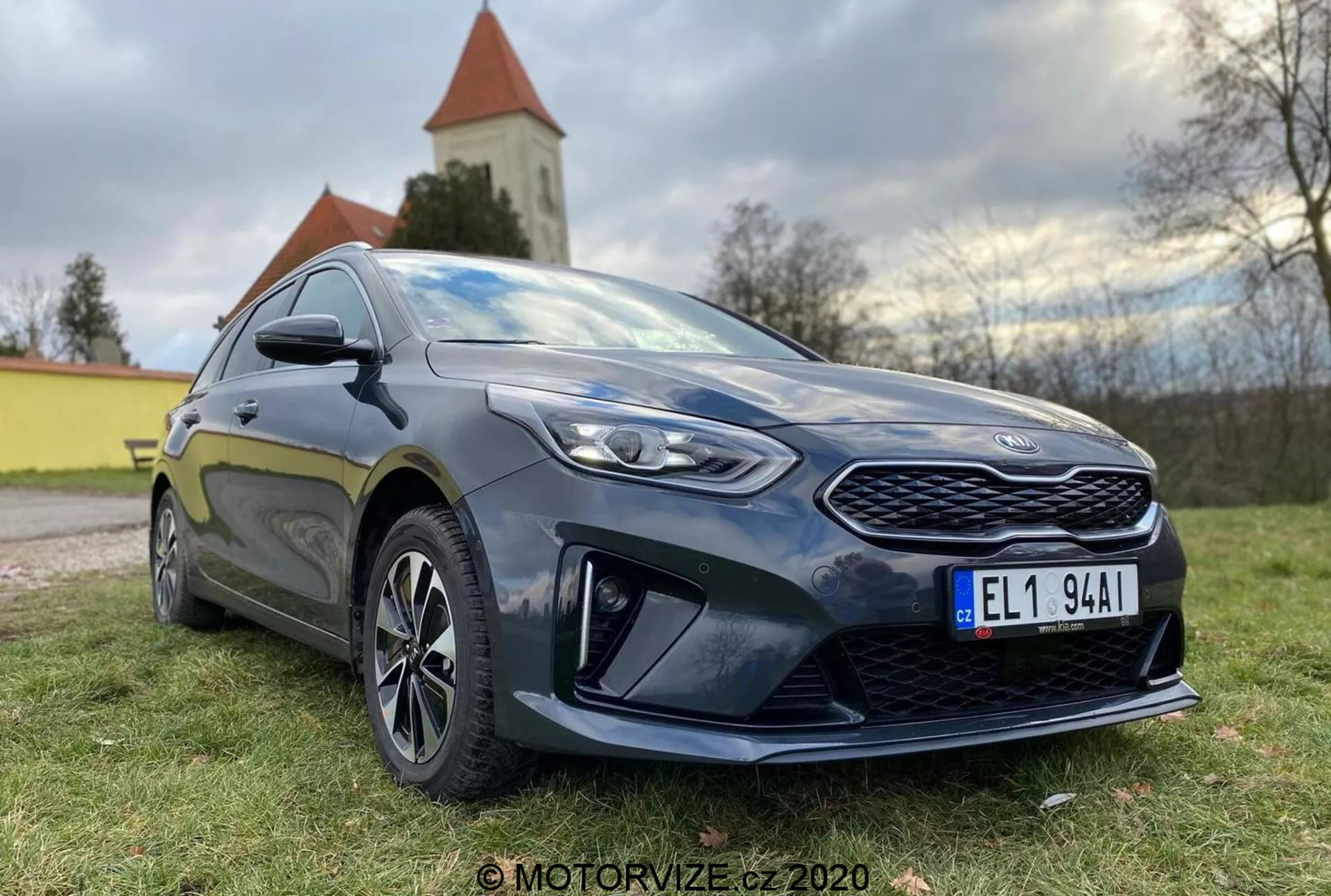 Vista detallada de un moderno hatchback Kia Ceed 2018, fotografiado desde un ángulo bajo de tres cuartos frontales, mostrando una parrilla prominente, faros LED angulares y el perfil lateral hasta la puerta del conductor. El coche es de color oscuro, probablemente azul oscuro o negro, con un brillo que resalta su estado bien mantenido. Los elementos de diseño notables incluyen una parrilla frontal con diseño de patrón, faros afilados con lentes claras, elementos aerodinámicos pronunciados en el parachoques delantero inferior, ruedas de aleación de varios radios, espejos laterales del color de la carrocería con indicadores integrados y un faldón lateral deportivo. La postura del coche sugiere un equilibrio entre comodidad y deportividad. La luz natural del día con iluminación suave resalta los detalles, y se puede ver un paisaje natural con una iglesia o torre en el fondo.