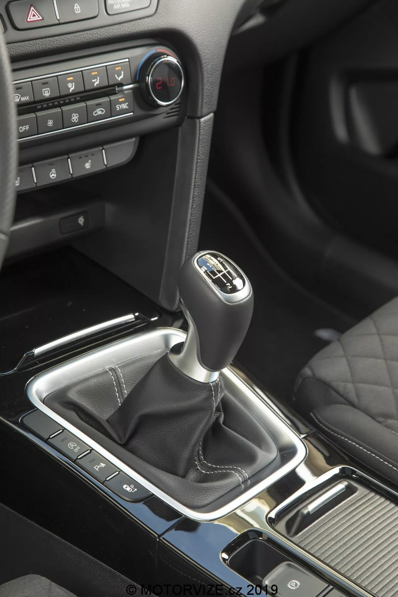 Vista cercana del área de cambio de marchas dentro de un Kia Ceed 2021 desde un ángulo ligeramente elevado, mostrando la palanca de cambios manual con un patrón de seis velocidades y una funda negra de material similar al cuero, consola central con acentos plateados de alto brillo, varios botones de control indicativos de funciones tecnológicas, elementos del tablero de superficie mate con botones de control climático, iluminación ambiental roja sutil, portavasos prácticos y mejoras estéticas texturizadas a la izquierda de la palanca de cambios.