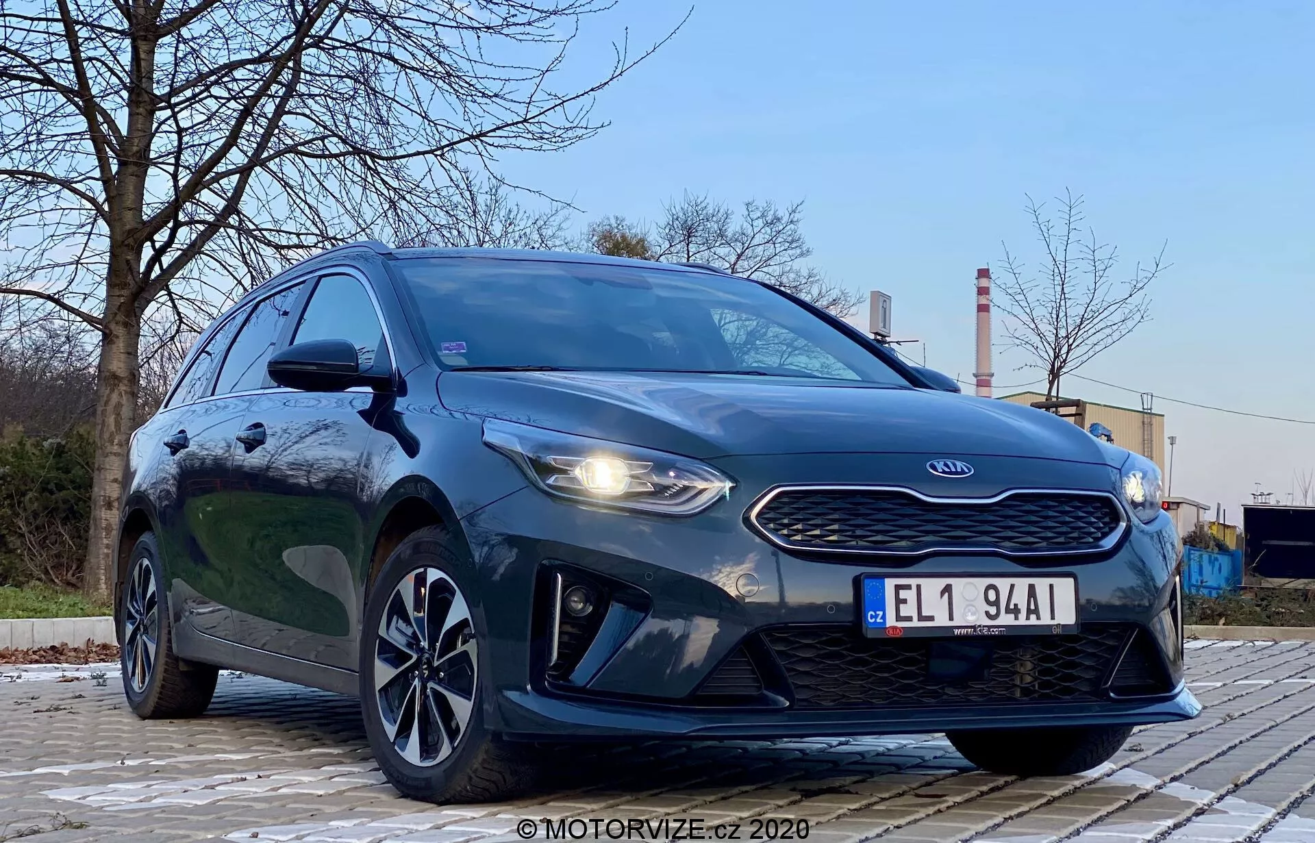 Fotografía del Kia Ceed 2018 tomada desde un ángulo bajo y delantero izquierdo, destacando la parrilla, los faros y el diseño de la fascia delantera del vehículo. El estilo agresivo del coche se resalta con los faros prominentes, la parrilla de nariz de tigre y los contornos del capó. La imagen también muestra las luces diurnas LED, el parachoques delantero aerodinámico y las elegantes llantas de aleación. El entorno es al aire libre con árboles y estructuras industriales de fondo, proporcionando contraste. La pintura metálica oscura del vehículo es brillante y bien iluminada, con reflejos que indican una superficie limpia. La imagen se centra en el coche con una perspectiva dramática de ángulo bajo bajo una suave iluminación exterior.