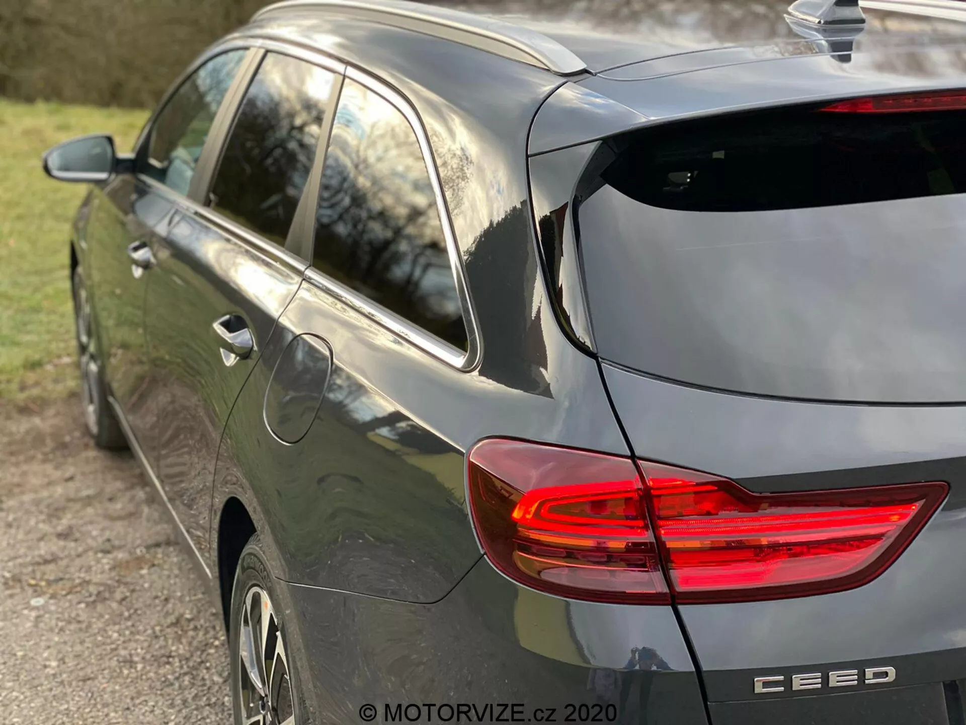 Vista parcial lateral y trasera de un moderno automóvil hatchback Kia Ceed 2018 de color gris, destacando la luz trasera LED en forma de C iluminada, diseño de hatchback con limpiaparabrisas trasero y pequeño alerón en el techo, antena de aleta de tiburón, ruedas de aleación y emblema de la marca con el nombre del modelo 'CEED', capturado con luz natural de tarde y poca profundidad de campo enfocándose en el vehículo.