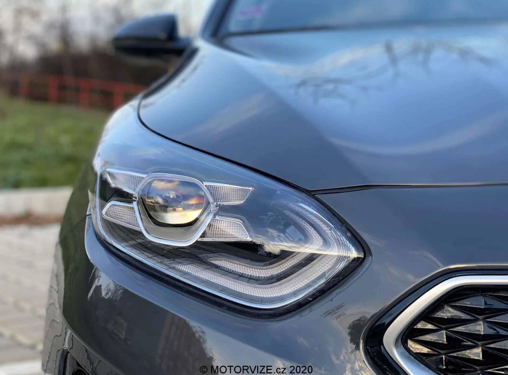 Vista detallada en primer plano de la parte delantera izquierda de un Kia Ceed 2018, centrada en su sofisticado faro y parte de la parrilla frontal. La foto es tomada desde un ángulo bajo, destacando el diseño elegante y moderno del coche con una tecnología de iluminación avanzada visible, posiblemente con faros LED y adaptativos. El fondo desenfocado acentúa los elementos aerodinámicos y la sensación de lujo del coche, indicando un vehículo orientado al rendimiento.
