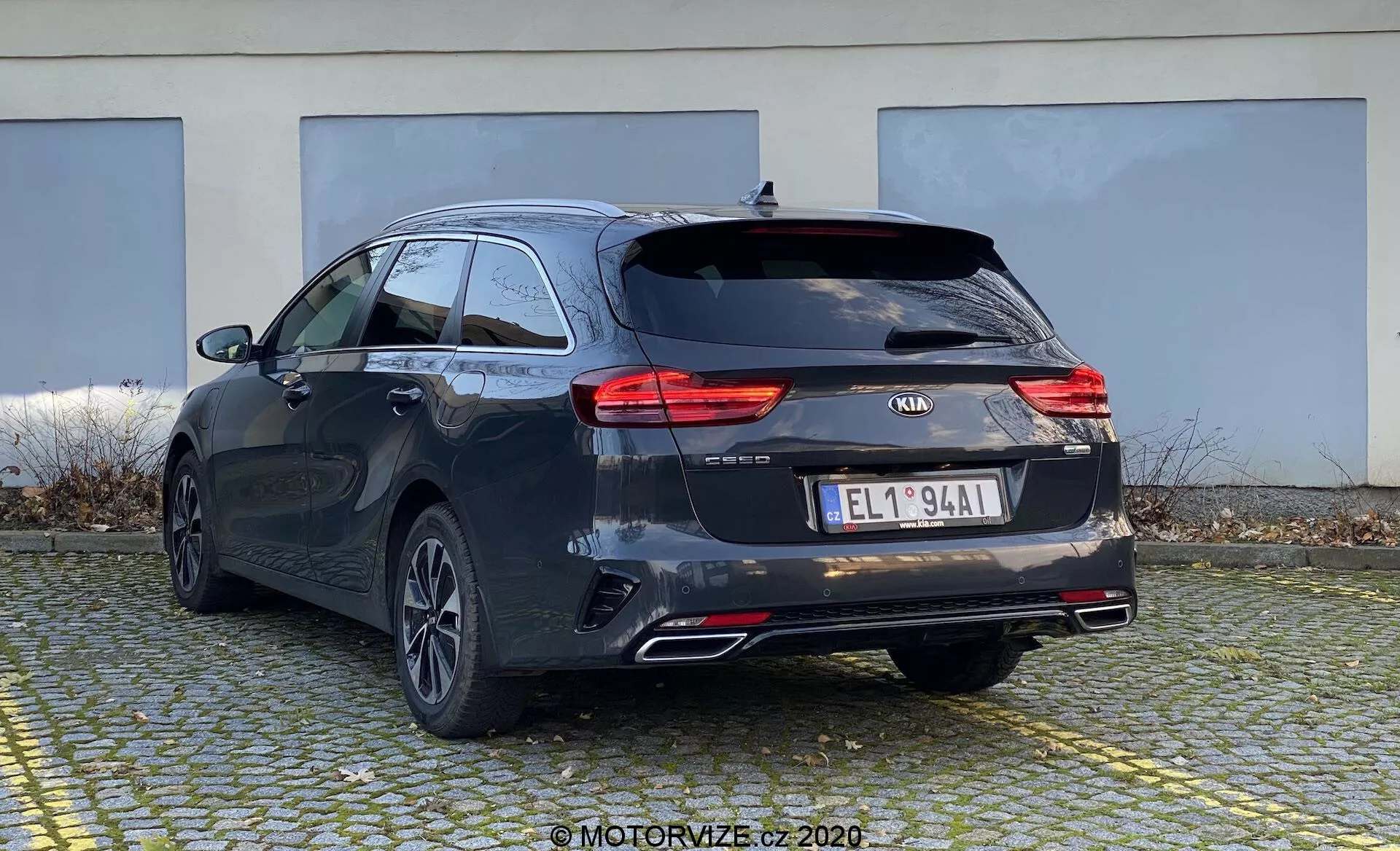 La imagen muestra un Kia Ceed 2018 desde una perspectiva trasera de tres cuartos, destacando la parte trasera y el lateral del coche familiar. El vehículo tiene un tono oscuro, posiblemente negro o gris oscuro, con líneas característicamente diseñadas y un techo ligeramente inclinado para un aspecto deportivo. Se observan luces traseras LED modernas, el logotipo de Kia, el nombre del modelo 'CEED', insignias adicionales, una matrícula de estilo europeo, llantas traseras de aleación con múltiples radios, doble tubo de escape y un limpiaparabrisas trasero. Probablemente tenga cámara trasera y sensores de estacionamiento que no son visibles. Un pequeño alerón de techo y rieles indican opciones adicionales de carga. El coche está estacionado en una zona pavimentada con un edificio al fondo, mostrando el perfil trasero y parte del lateral desde un ángulo bajo.