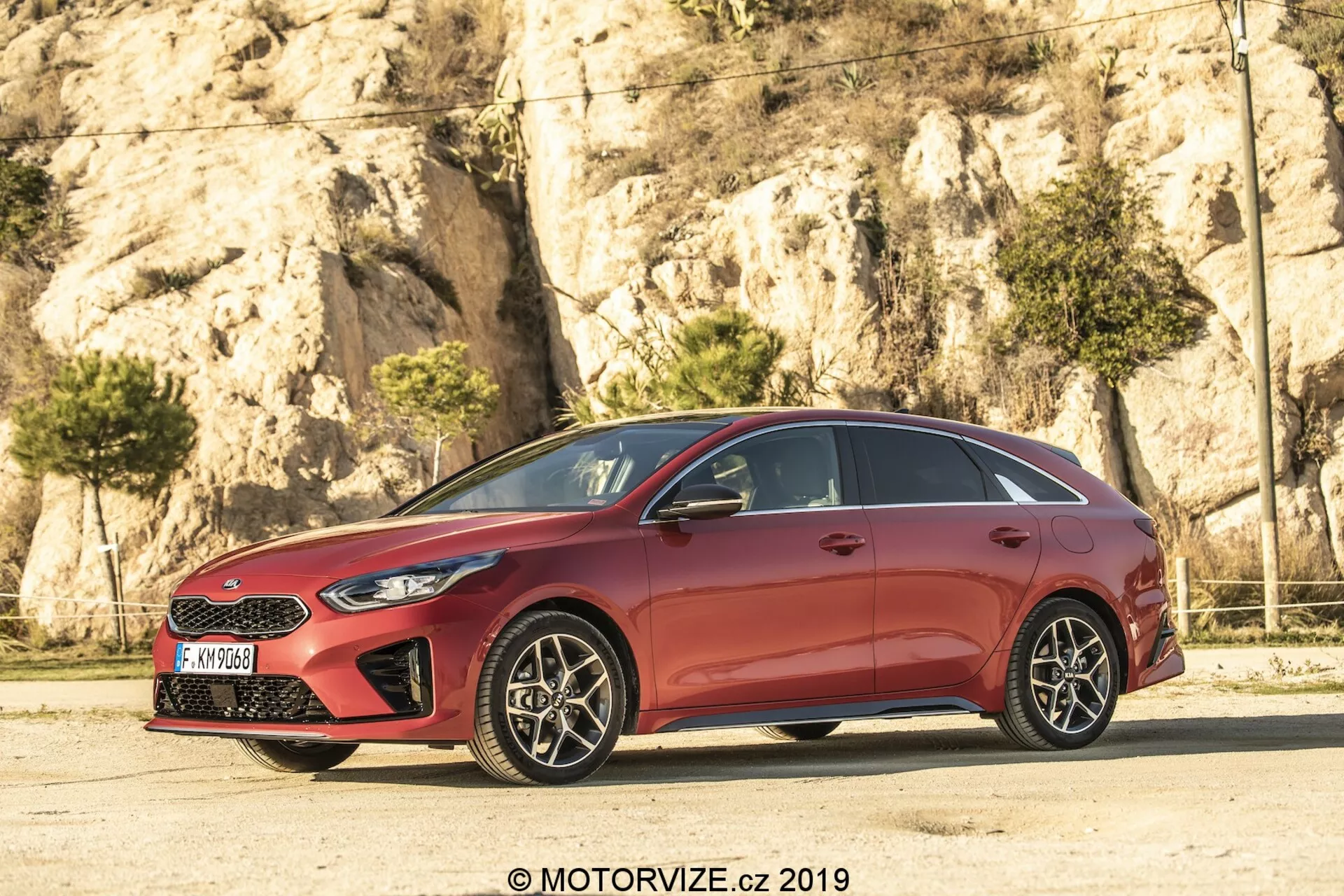 Vista de perfil lateral de un hatchback Kia Ceed rojo 2021, con un diseño elegante y aerodinámico, líneas de techo inclinadas y líneas contorneadas en las puertas. El coche, de cinco puertas, tiene molduras cromadas en las ventanas y pilares B en negro, creando un efecto de panel de vidrio continuo. Las llantas de aleación de múltiples radios, con frenos de disco visibles, agregan un toque deportivo. Los faros afilados y la parrilla parcialmente visible marcan el frente, mientras que los espejos laterales del color del cuerpo con señales de giro integradas mejoran la visibilidad y la seguridad.