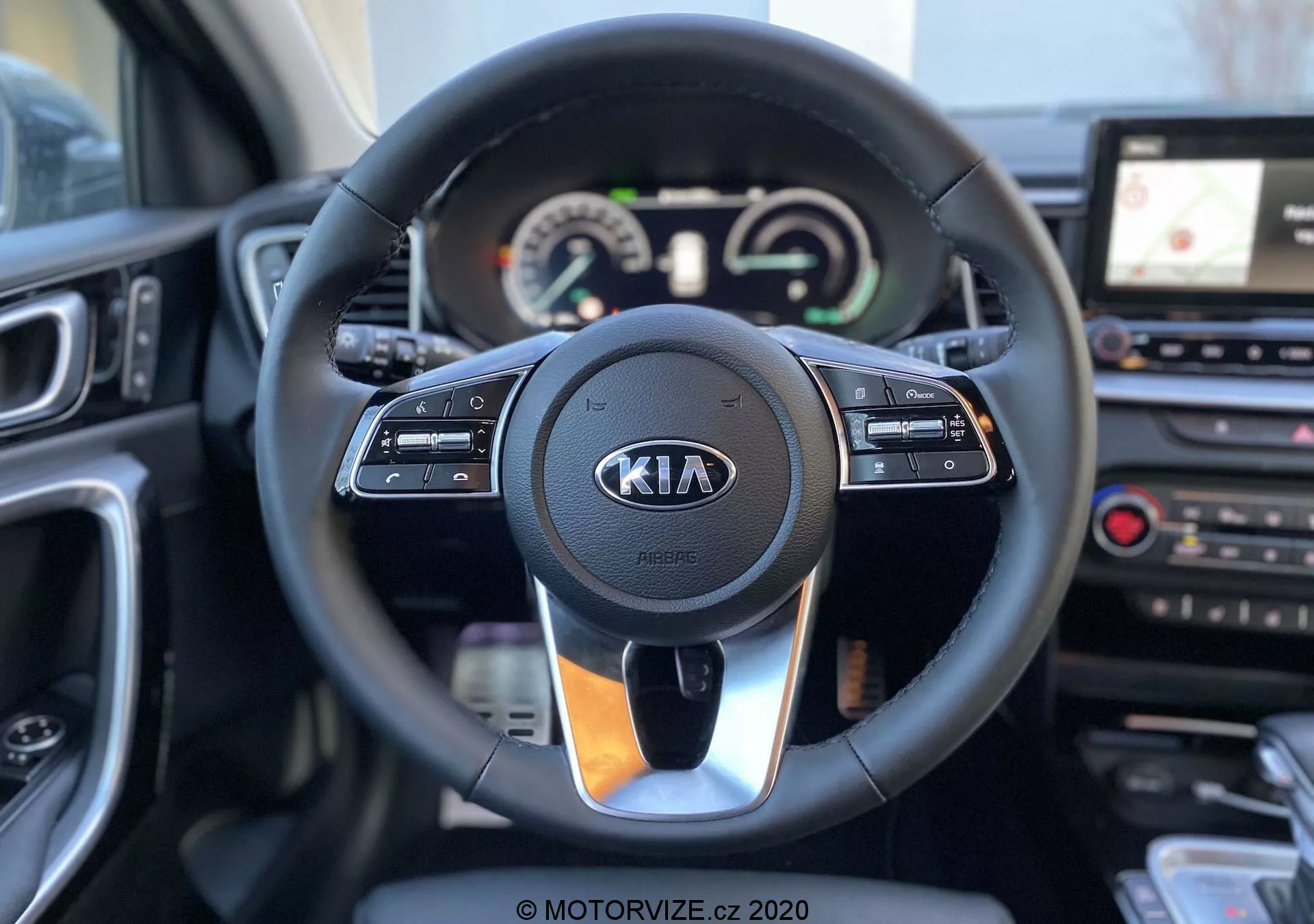 Vista interior de un Kia Ceed 2018 centrada en el volante y el tablero de instrumentos desde la perspectiva del conductor. El moderno volante cuenta con controles para el control de crucero, interacción multimedia y comunicación manos libres, con el logo de Kia claramente visible. Detrás del volante, un cuadro de instrumentos combina diales analógicos y una pantalla digital para la lectura del velocímetro y el tacómetro, junto a una pantalla multifuncional. A la derecha del volante, se puede ver parte de la consola central que incluye un sistema de infoentretenimiento con pantalla táctil y navegación GPS, así como botones de control de climatización. La imagen es clara con una profundidad de campo que resalta el volante y sus alrededores inmediatos.
