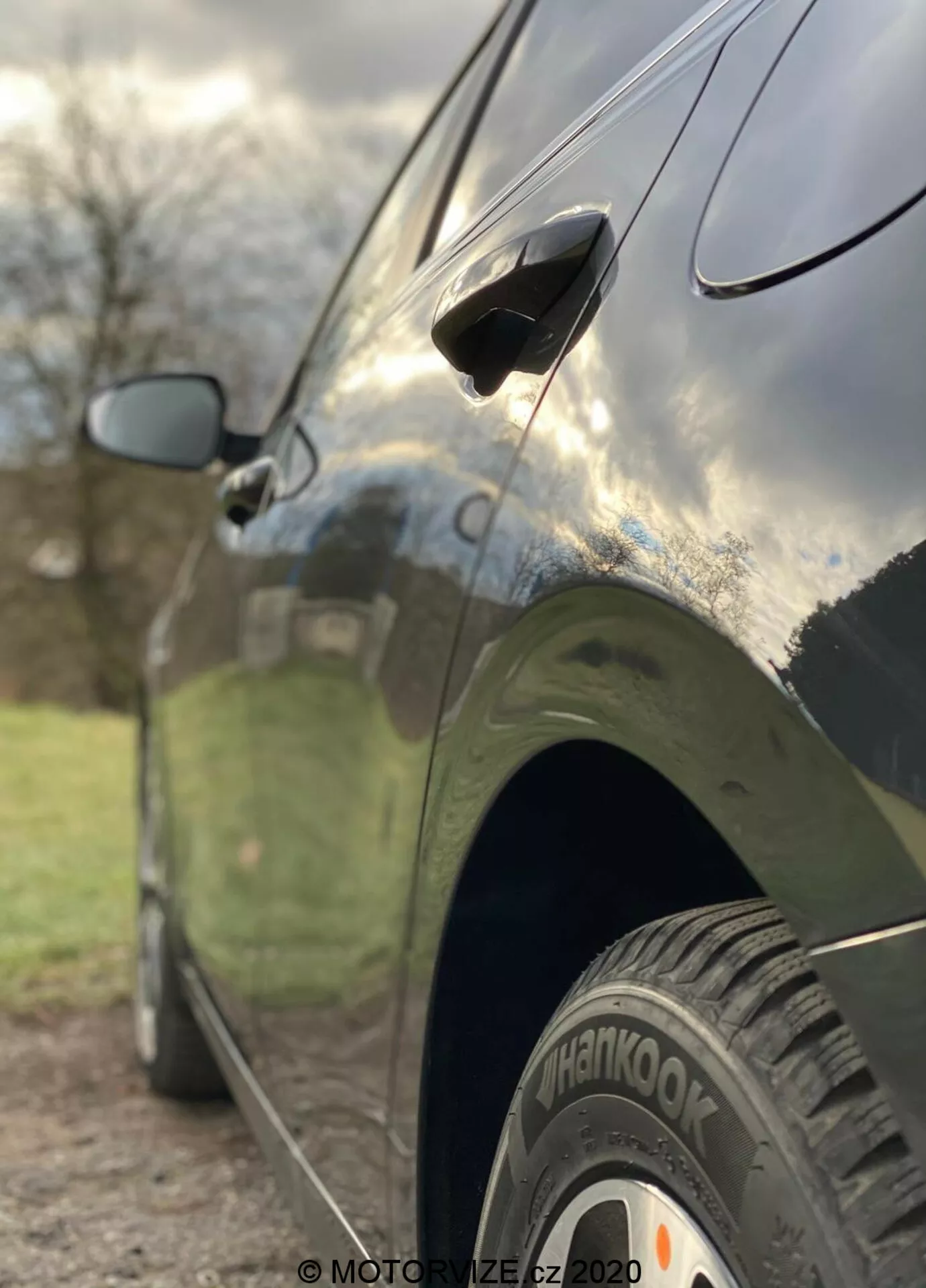 Vista lateral en primer plano del Kia Ceed 2018, destacando características como la rueda trasera con neumático 'Hankook', manija de puerta aerodinámica y espejo lateral parcialmente desenfocado. La foto está tomada desde un ángulo bajo con una profundidad de campo reducida que enfoca nítidamente la rueda trasera y la parte inferior de la puerta, mientras que el fondo está artísticamente desenfocado. El coche tiene un acabado oscuro y brillante que refleja el cielo y los alrededores, mostrando líneas de diseño elegantes y una carrocería bien mantenida.