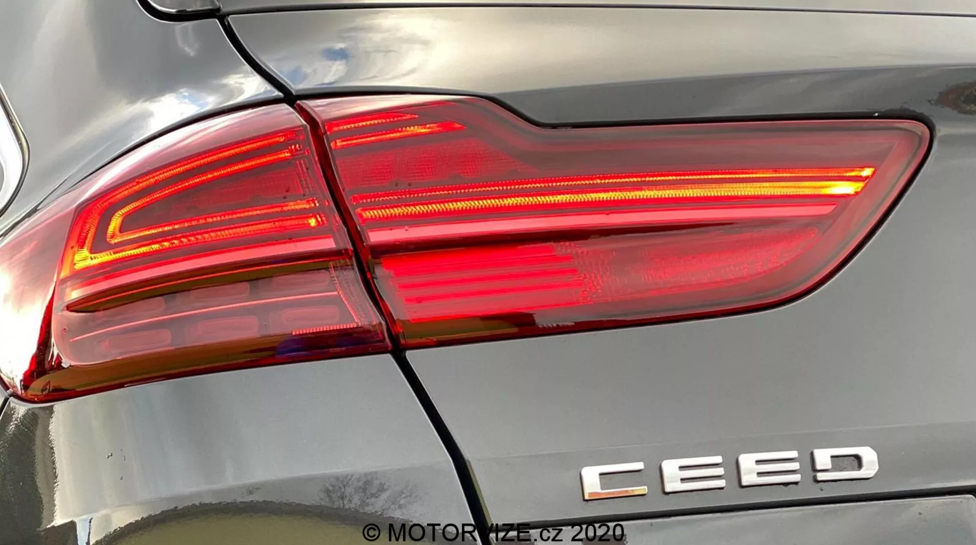 Vista detallada en primer plano del conjunto del faro trasero derecho de un Kia Ceed 2018, iluminado, mostrando un diseño moderno con líneas angulares afiladas, secciones de lentes claras y rojas, y la insignia del modelo 'CEED'. Una línea horizontal separa el faro trasero del parachoques trasero. Los reflejos en la carrocería destacan la curvatura de la superficie. Marca de agua 'MOTOR1.cz ©2020'.