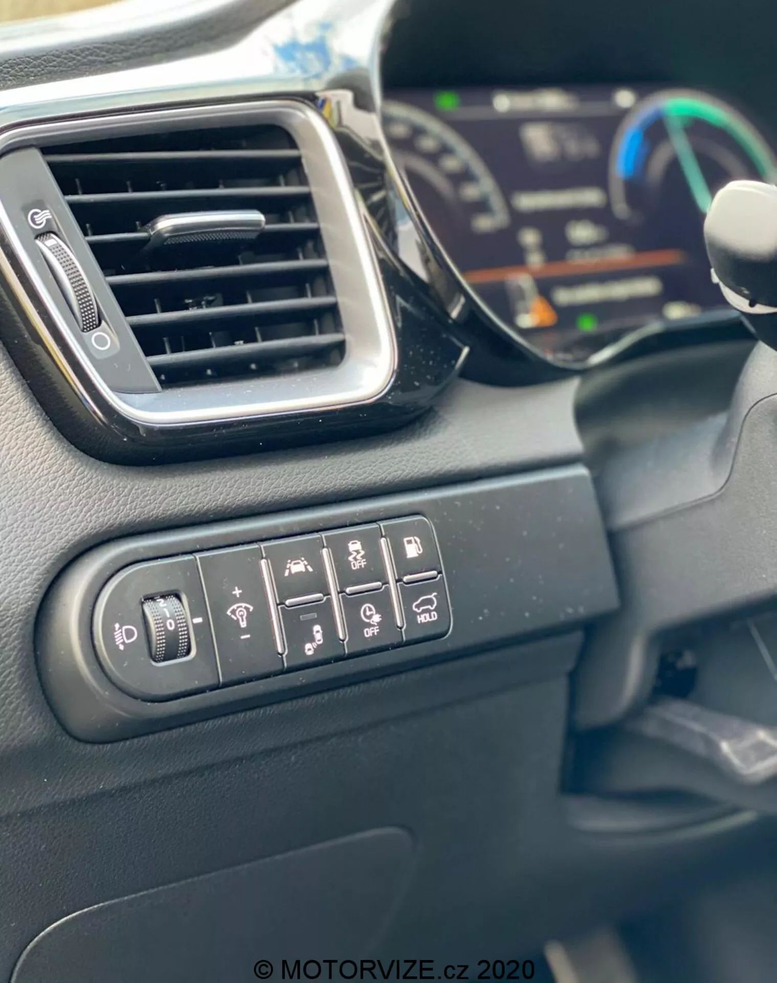 Vista en primer plano del interior del Kia Ceed 2018 que muestra los botones de control para ajuste de espejos laterales, calefacción de la ventana trasera, calefacción del volante, advertencia de salida de carril, asistencia de aparcamiento y control de tracción. La imagen también presenta una ventila de aire elegante, parte de un panel de instrumentos digital en el fondo borroso y el borde del volante. Los materiales interiores son principalmente plásticos duros con acabado mate, capturados a la luz del día.