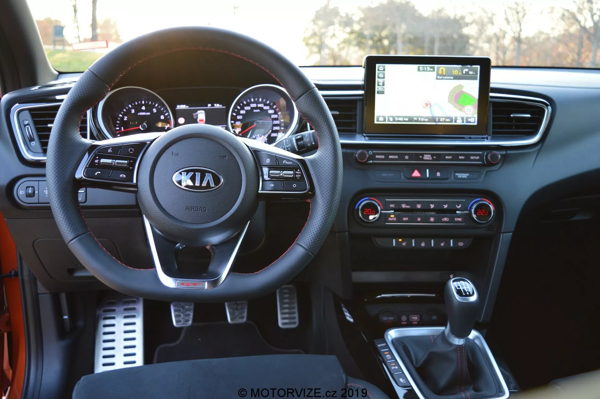 Vista interior de un Kia Ceed 2021 desde la perspectiva del conductor, mostrando el detallado tablero. La imagen muestra prominentemente el volante con el logo de Kia y controles incorporados para audio, llamadas y control de crucero. Detrás de él, se puede ver el cuadro de instrumentos con indicadores analógicos acentuados con cromo y una pantalla digital. La pantalla táctil central de infoentretenimiento muestra la navegación GPS, mientras que debajo están los botones y perillas de los controles de clima. La consola central muestra la palanca de cambios, portavasos y un puerto USB para la conectividad de dispositivos, destacando la tecnología moderna en el automóvil.