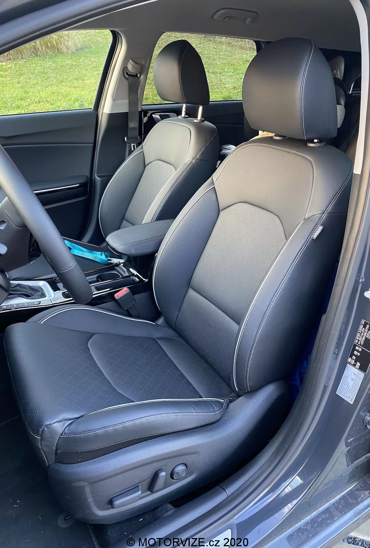 Cercano del interior de un Kia Ceed 2018, mostrando los asientos del conductor y del pasajero con tapicería de cuero bitono y costuras contrastantes, diseño de asiento tipo bucket con refuerzos laterales, y ajustes eléctricos en el asiento del conductor para el control del respaldo y la posición. Pliegues ligeros indican uso y hay varios artículos visibles en el asiento del pasajero.