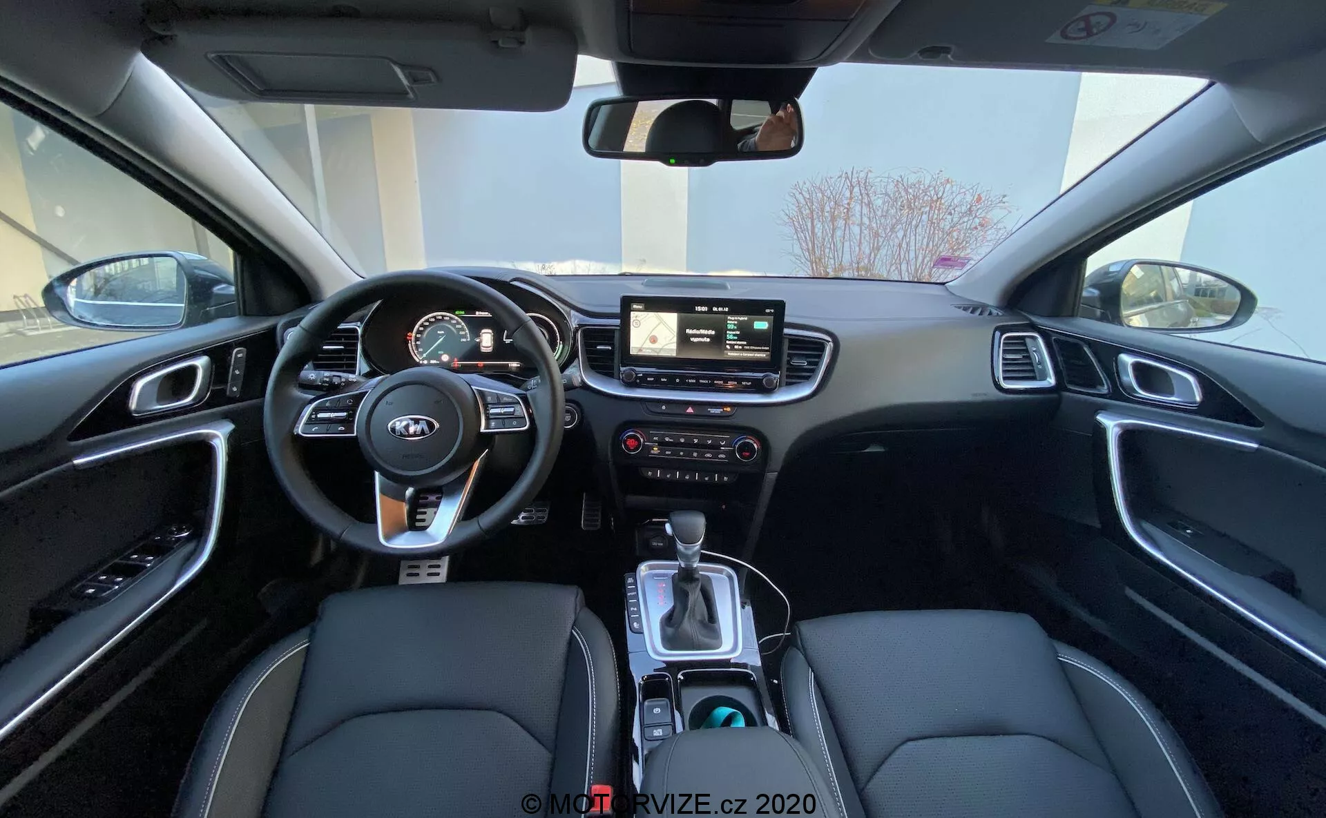 Vista gran angular del interior del Kia Ceed 2018 desde el asiento trasero, que muestra el área del conductor, incluido el volante con controles montados, el panel de instrumentos con medidores digitales y analógicos, el sistema de infoentretenimiento central con pantalla táctil, controles manuales para el infotainment y el clima, y la consola central con palanca de cambios, selector de modo de conducción, portavasos y un interior negro que presenta asientos de cuero sintético con costuras blancas y molduras de aluminio cepillado.