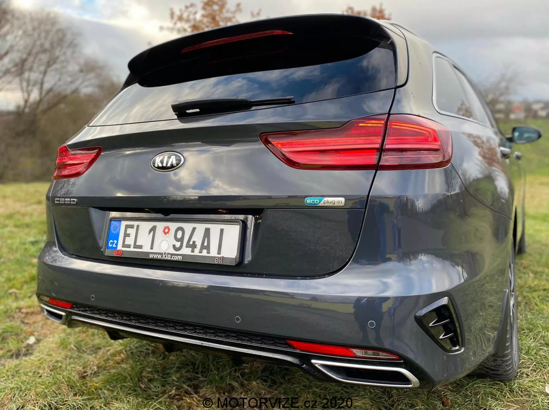 Vista trasera de un Kia Ceed Proceed 2018 de color gris oscuro, con características detalladas como luces traseras LED horizontales, un spoiler trasero montado en el techo, tubos de escape dobles cromados, parachoques trasero de estilo difusor negro y distintivos centrales que incluyen 'Kia' y 'GT Line.' La cámara de visión trasera sobre la matrícula central sugiere asistencia de aparcamiento, mientras que la poca profundidad de campo enfatiza la marca y los identificadores del modelo del coche en un fondo de césped.
