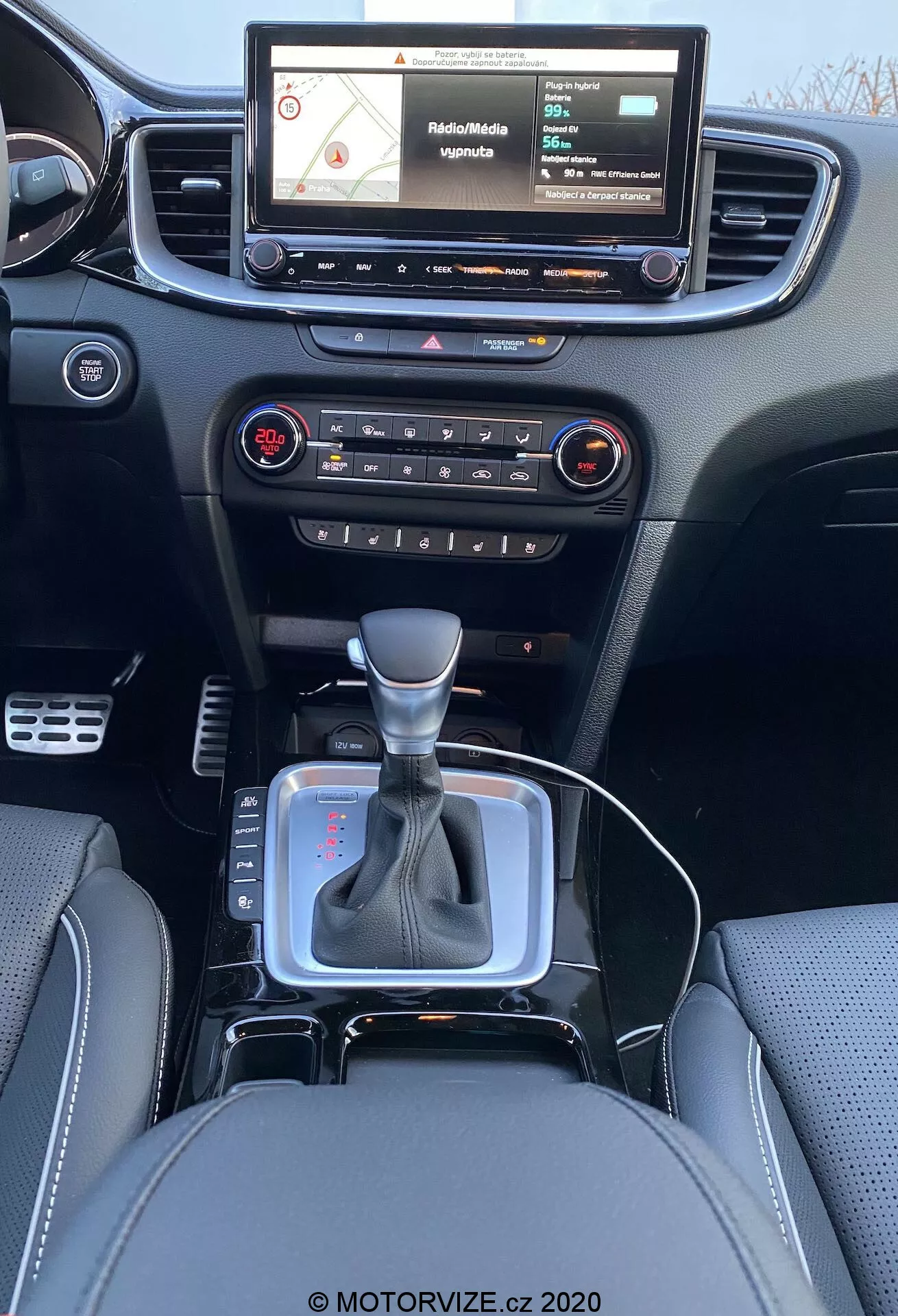 Vista interior de un Kia Ceed 2018 desde la perspectiva del asiento trasero, mostrando un moderno salpicadero con un sistema de infoentretenimiento con pantalla táctil que muestra un mapa de navegación, botones físicos y perillas para el control de audio y clima, ventilación central, palanca de cambios en posición neutral con acento plateado y funda de cuero negro, pequeño espacio de almacenamiento cerca de la palanca de cambios posiblemente para un enchufe de 12V o puertos USB, asientos de cuero con costuras detalladas.