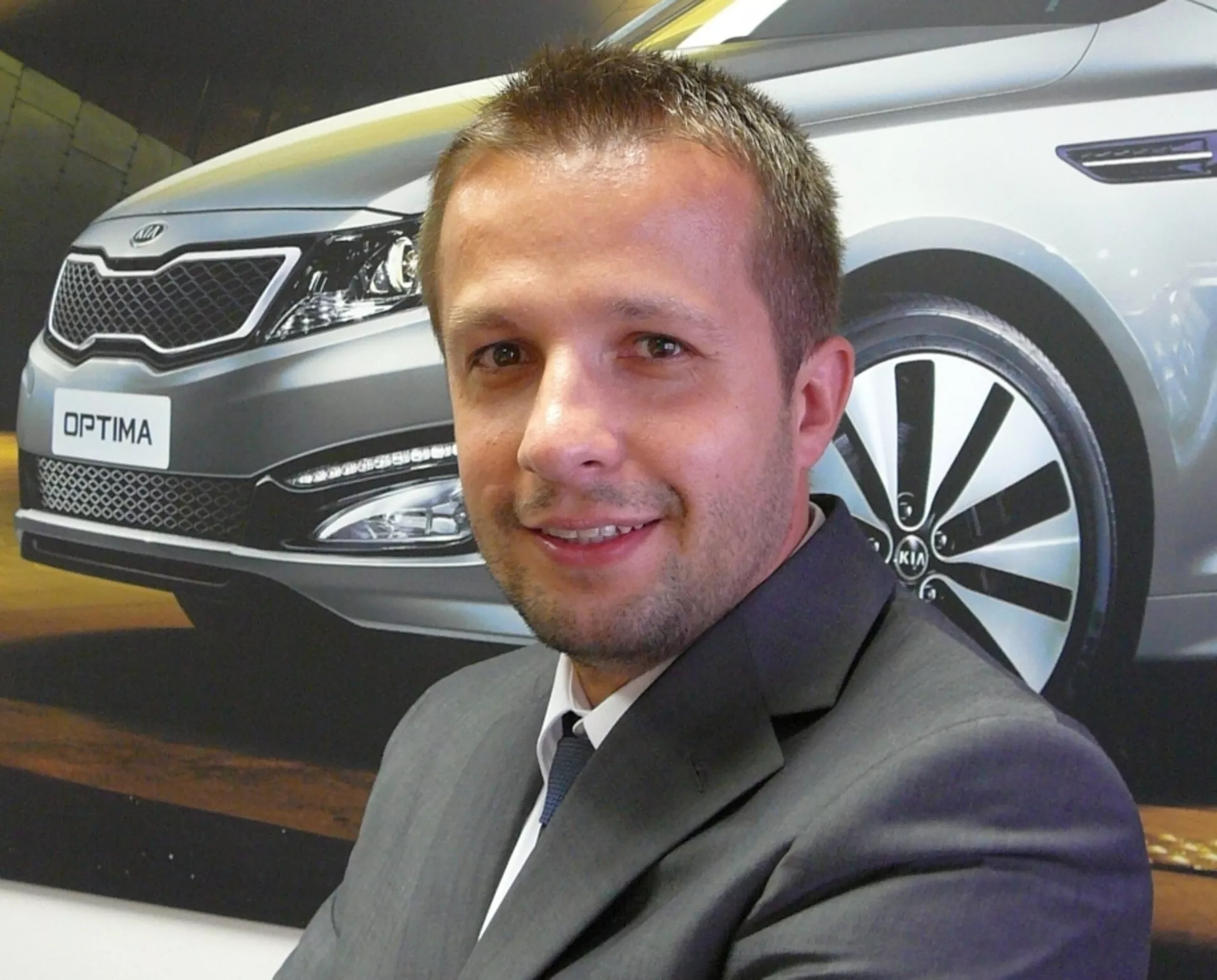 Hombre posando delante de un póster promocional de Kia Optima, mostrando la vista de cuarto frontal del coche con faros LED, parrilla cromada y diseño de rueda de aleación.