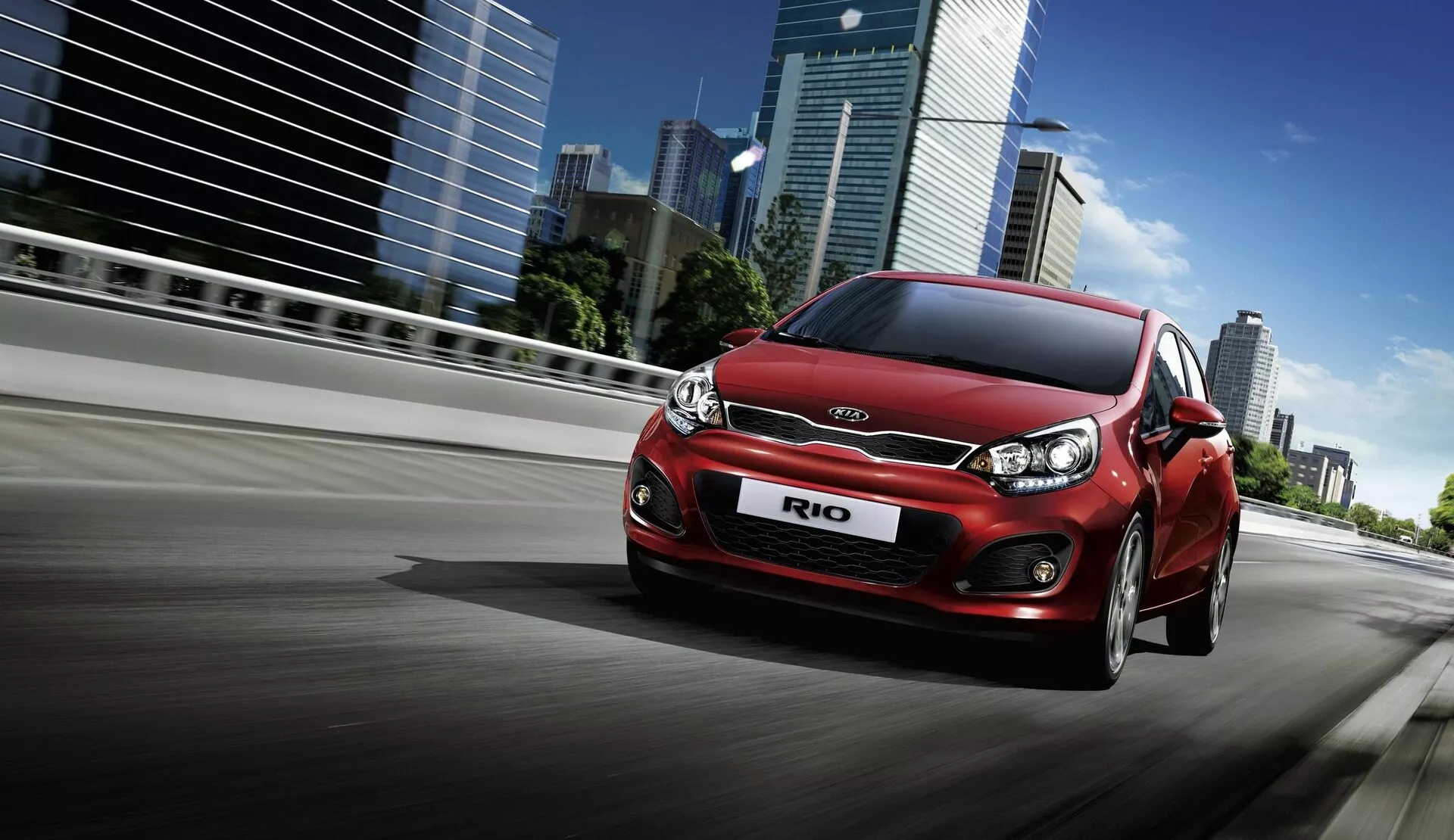 Hatchback rojo Kia Rio 2005 en movimiento en una carretera urbana con edificios altos de fondo, mostrando su diseño desde una vista frontal tres cuartos baja.