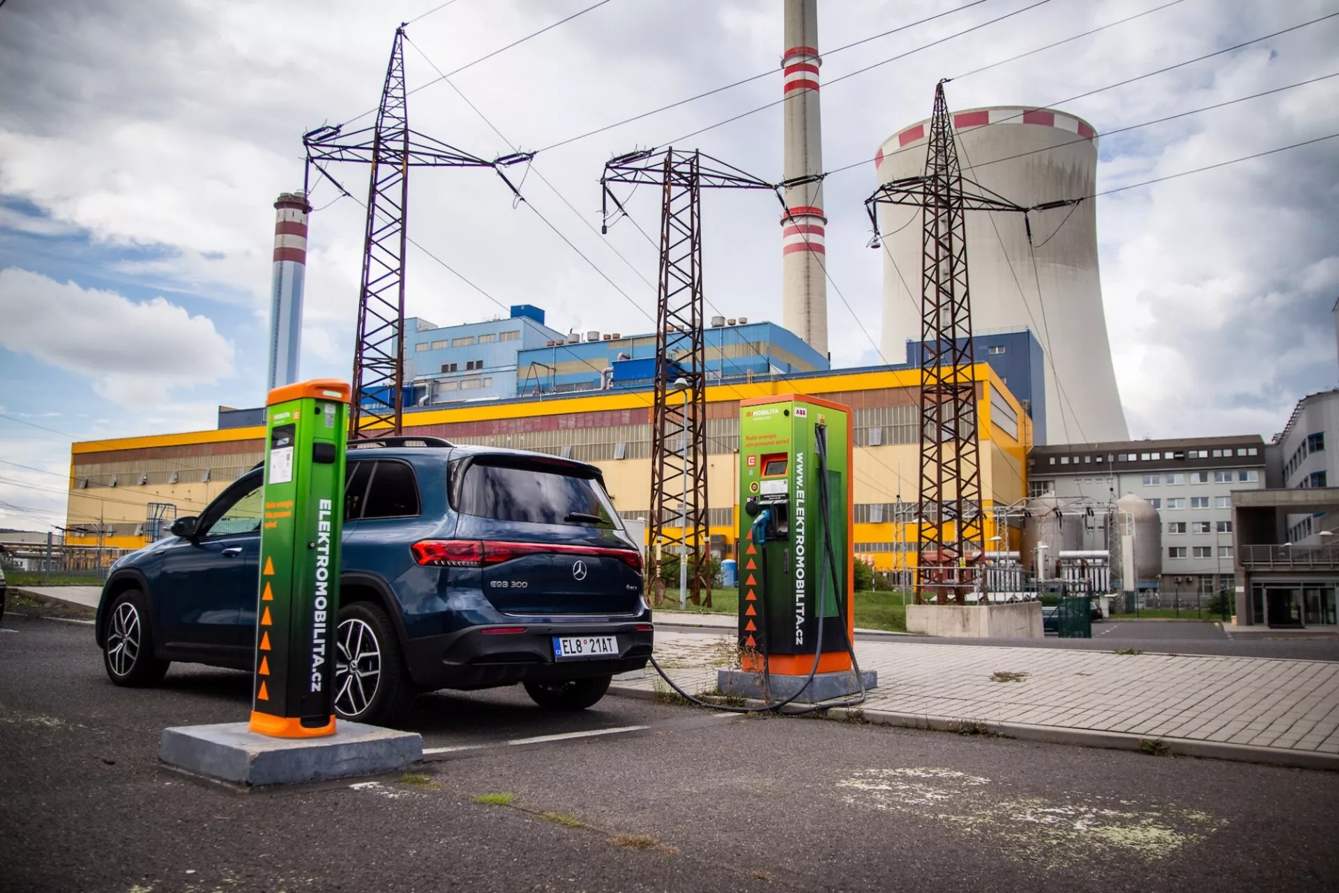 Hatchback eléctrico Mercedes-Benz EQB de 2021 estacionado en una estación de carga con un cable enchufado, visto desde la parte trasera tres cuartos, destacando sus luces traseras, ventana y lado del pasajero contra un fondo industrial
