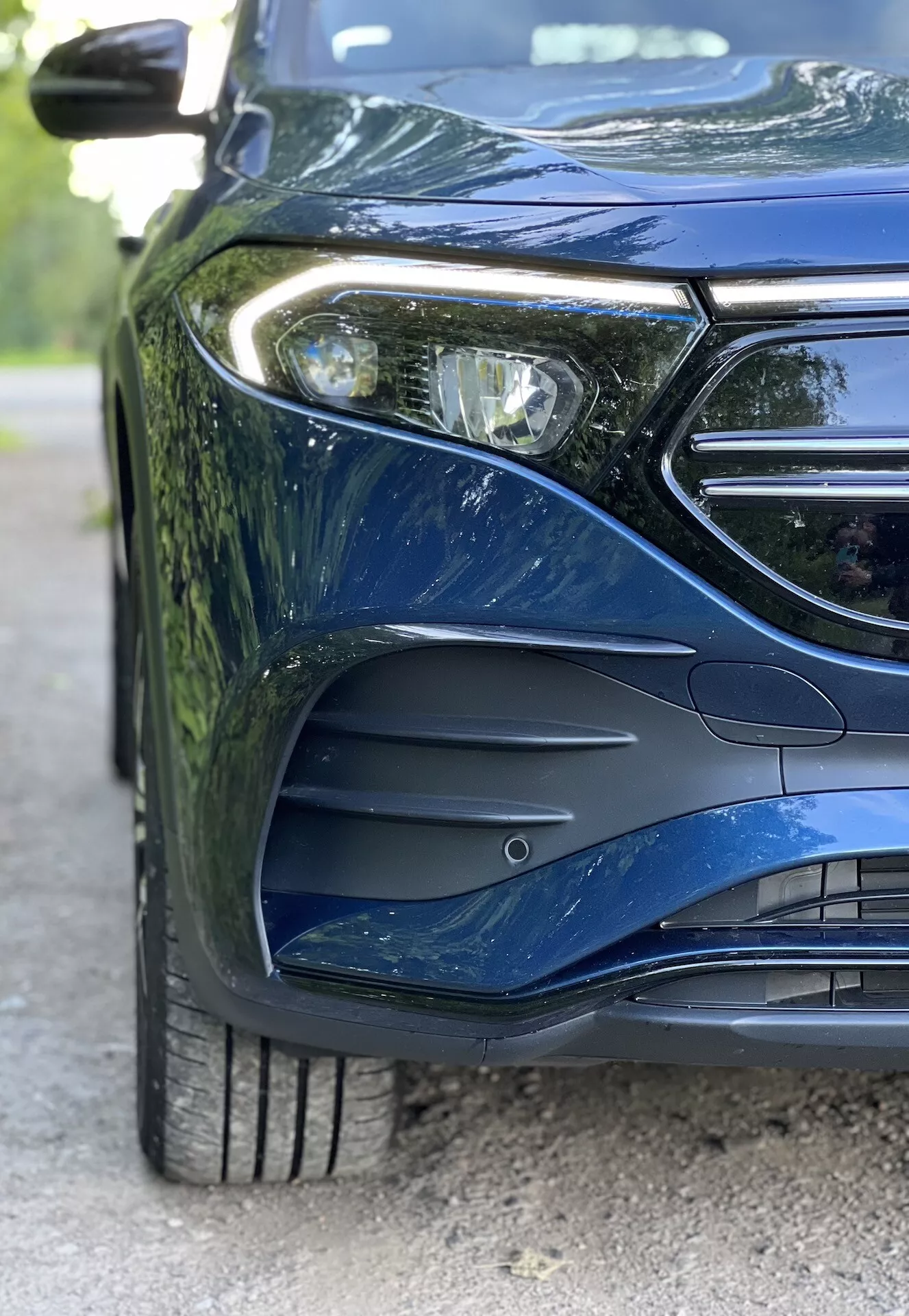Vista de cerca del lado frontal izquierdo del Mercedes-Benz EQB 2021, mostrando el diseño del faro LED, parte de la parrilla con espacio para el emblema, parachoques frontal aerodinámicamente esculpido con entradas de aire, rueda de aleación de múltiples radios con dibujo detallado del neumático y pintura metálica que refleja curvas sofisticadas del cuerpo
