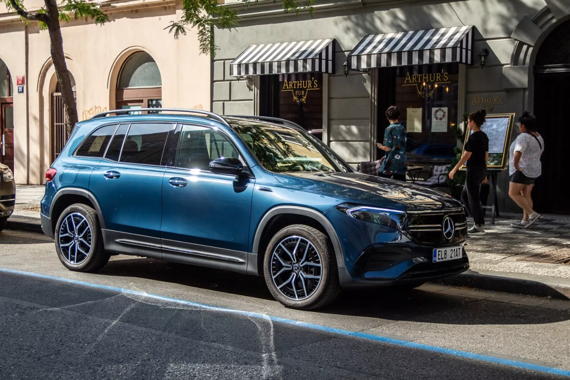 Perfil lateral del Mercedes-Benz EQB 2021 en la calle de la ciudad, mostrando el diseño moderno de SUV de lujo con grandes ruedas de aleación, señales de giro integradas en los espejos laterales, ventanas tintadas reflejando el entorno urbano, líneas de carrocería elegantes, rieles de techo y manijas de puerta al ras en iluminación natural