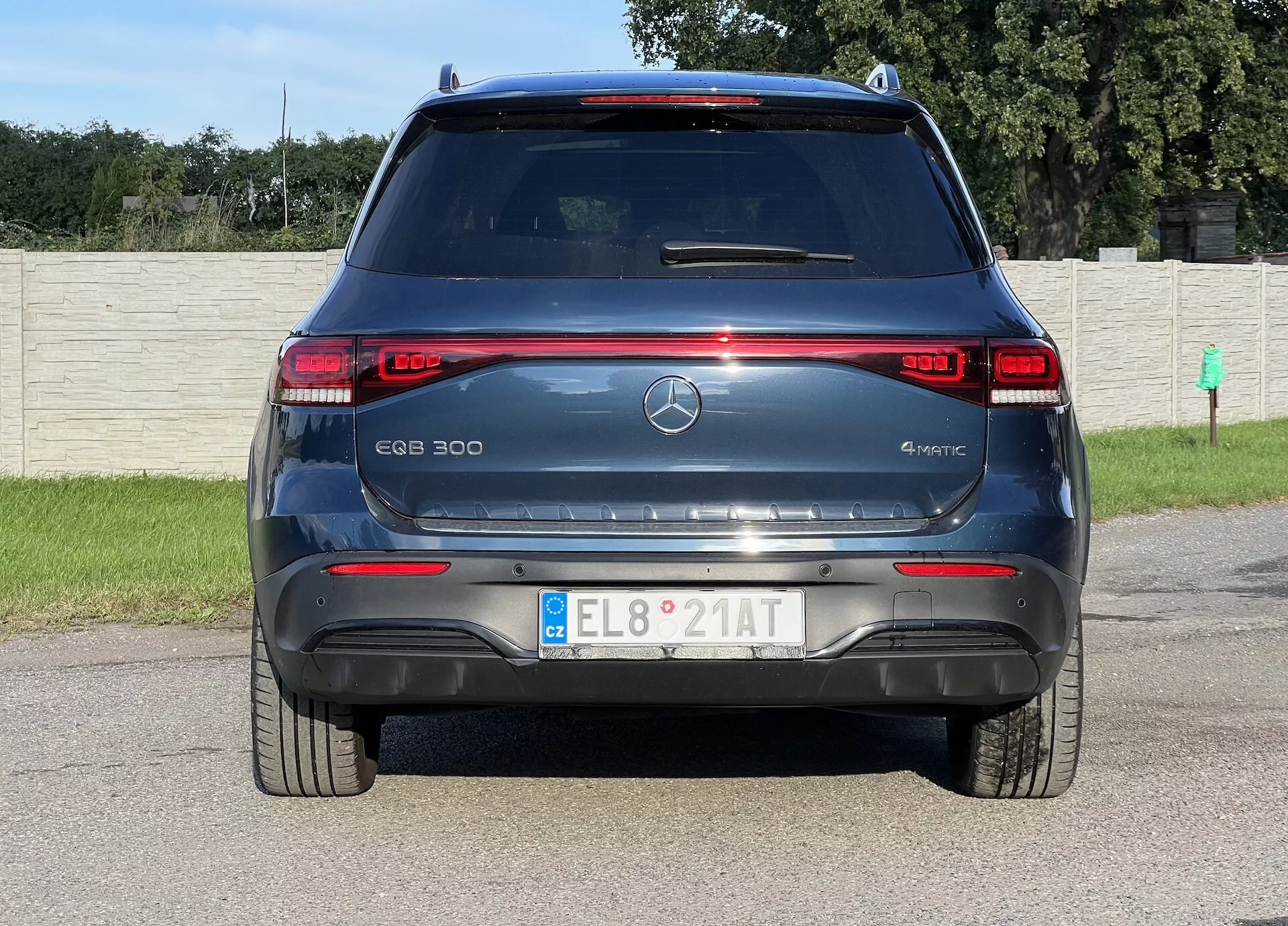 Vista trasera del SUV eléctrico Mercedes-Benz EQB 300 4MATIC mostrando luces traseras LED, marca del modelo y de electricidad, emblema de estrella de tres puntas, parabrisas trasero con desempañador y limpiaparabrisas, sensores de estacionamiento, reflectores rojos, placa de matrícula europea, spoiler trasero aerodinámico y sistema de tracción total 4MATIC, contra un fondo de campo de hierba y valla.