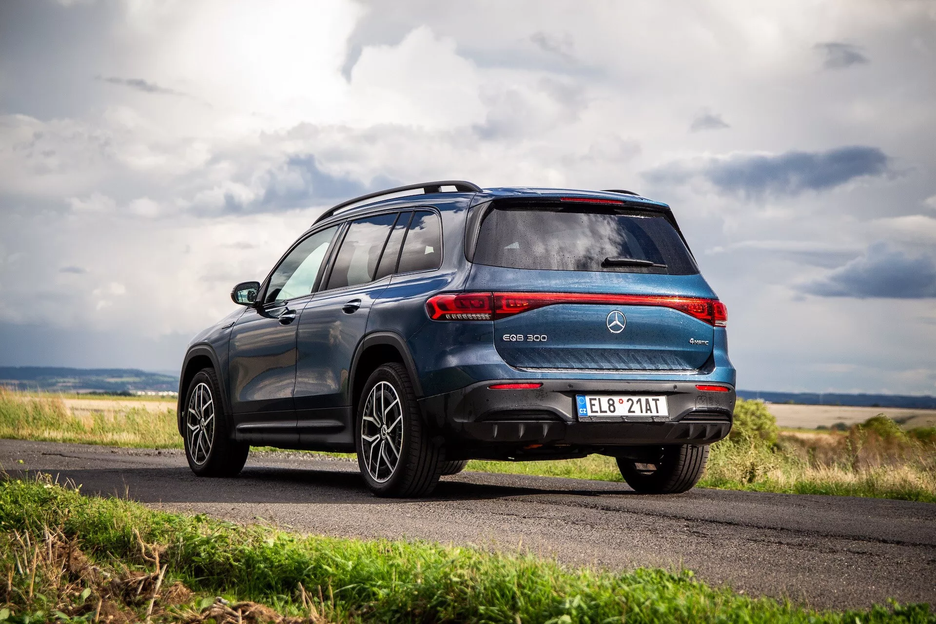 SUV eléctrico Mercedes-Benz EQB 2021 mostrada desde un ángulo trasero dinámico de tres cuartos demostrando el elegante diseño del vehículo, que incluye lámparas combinadas traseras, un alerón trasero deportivo, ruedas de aleación y distintivo EQB contra un telón de fondo de cielo nublado y paisaje rural.