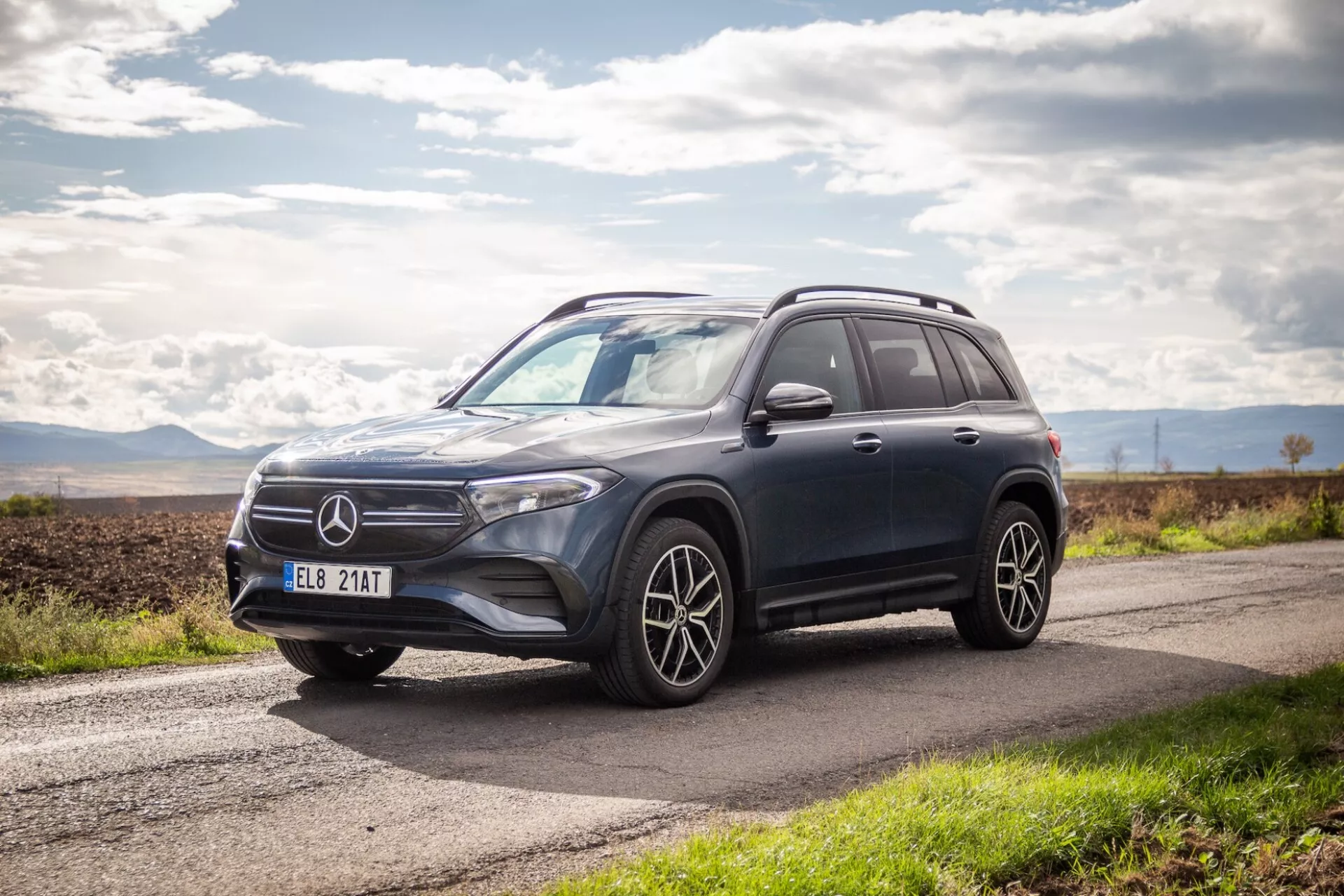 Vista lateral y frontal del SUV de lujo Mercedes-Benz EQB 2021 mostrando un diseño aerodinámico, faros LED, ruedas de aleación de múltiples radios y alta distancia al suelo en una carretera de asfalto bajo un cielo parcialmente nublado