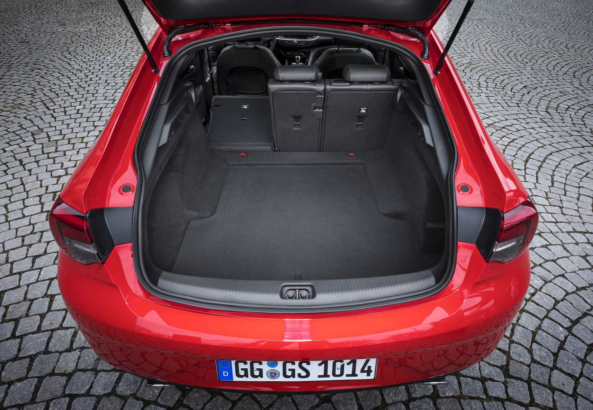 Vista trasera de un Opel Insignia rojo del 2013 con el maletero abierto, mostrando un área de carga espaciosa con revestimiento de moqueta, puntos de anclaje para almacenamiento seguro y un parachoques trasero con placa protectora negra, bajo iluminación ambiental