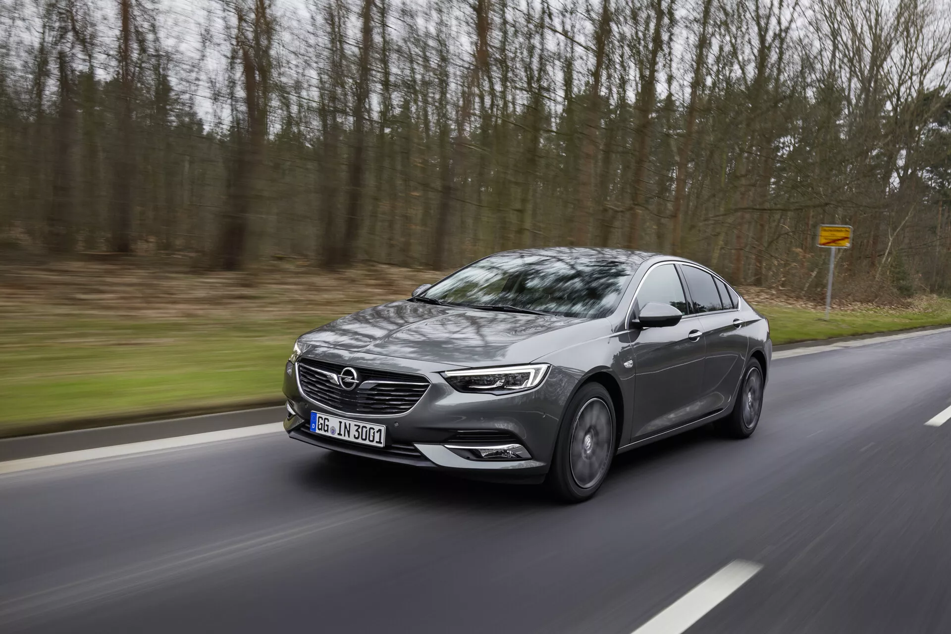 Sedán moderno Opel Insignia 2013 en movimiento por un camino sinuoso a través del bosque, mostrando perfil lateral con faros LED, parrilla grande, llantas de aleación, espejos laterales con indicadores y ventanas tintadas