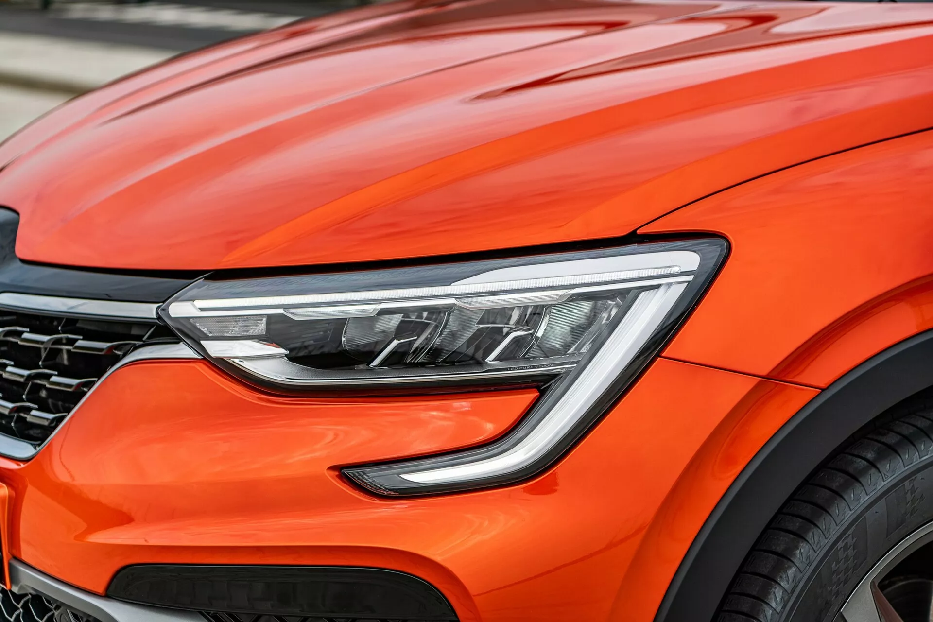 Vista de cerca de la parte frontal del Renault Arkana 2021 de color naranja brillante, enfocada en los detalles de diseño modernos como los faros LED avanzados, las curvas aerodinámicas del capó, una parrilla con diseño de panal parcialmente visible y un fragmento de una rueda de aleación grande, sugiriendo un rendimiento premium y tecnología automotriz de vanguardia.