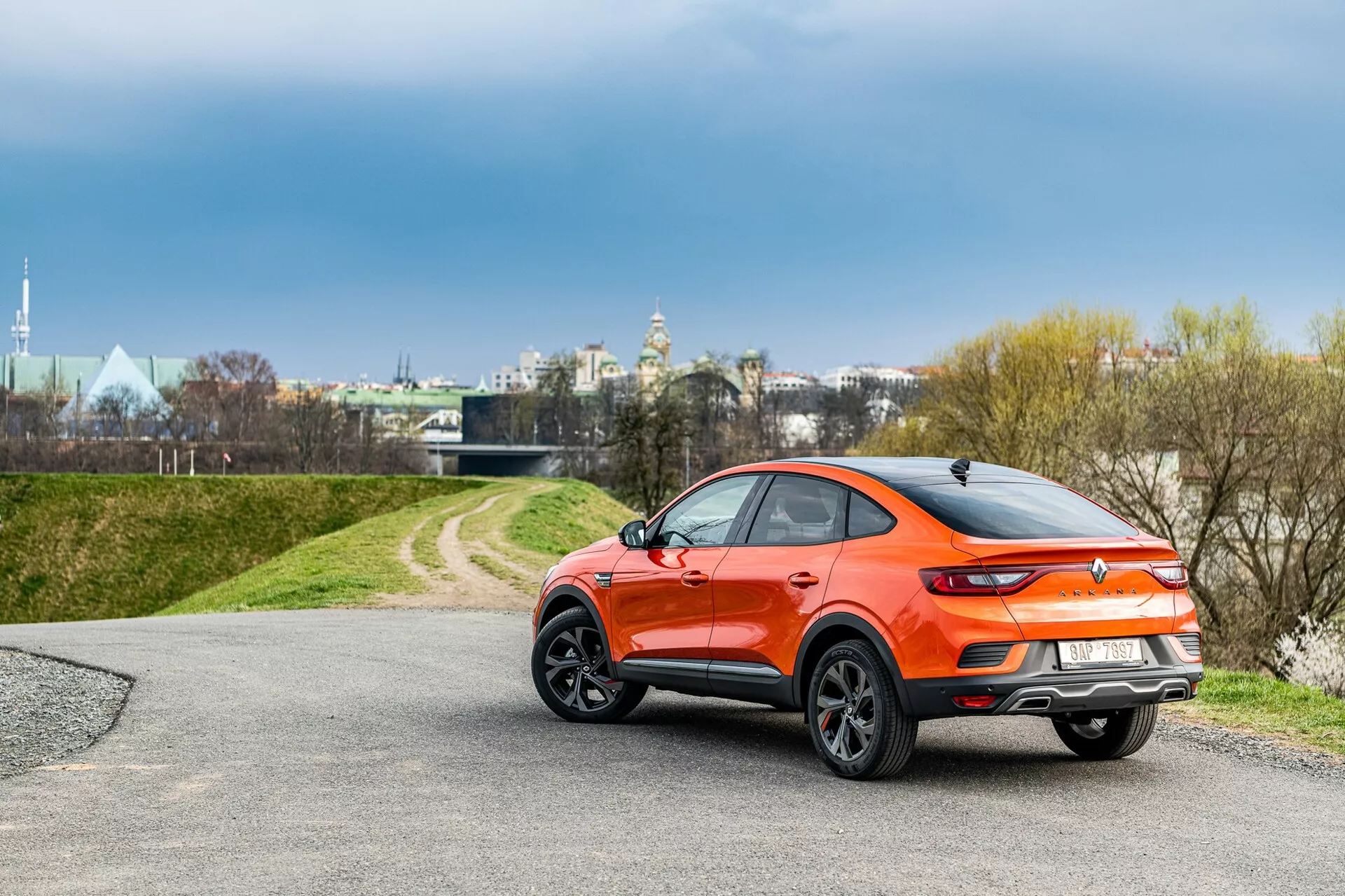 Vista trasera de tres cuartos de un Renault Arkana hatchback 2021 con diseño crossover deportivo en pintura metálica naranja vibrante. El coche presenta una línea de techo inclinada, molduras de plástico negro alrededor de los arcos de las ruedas, un alerón en la parte superior del portón trasero, luces traseras LED elegantes y grandes llantas de aleación multirradio. Está posicionado en una carretera asfaltada que conduce a un área de césped, bajo condiciones de cielo nublado cerca de un entorno urbano pero tranquilo.