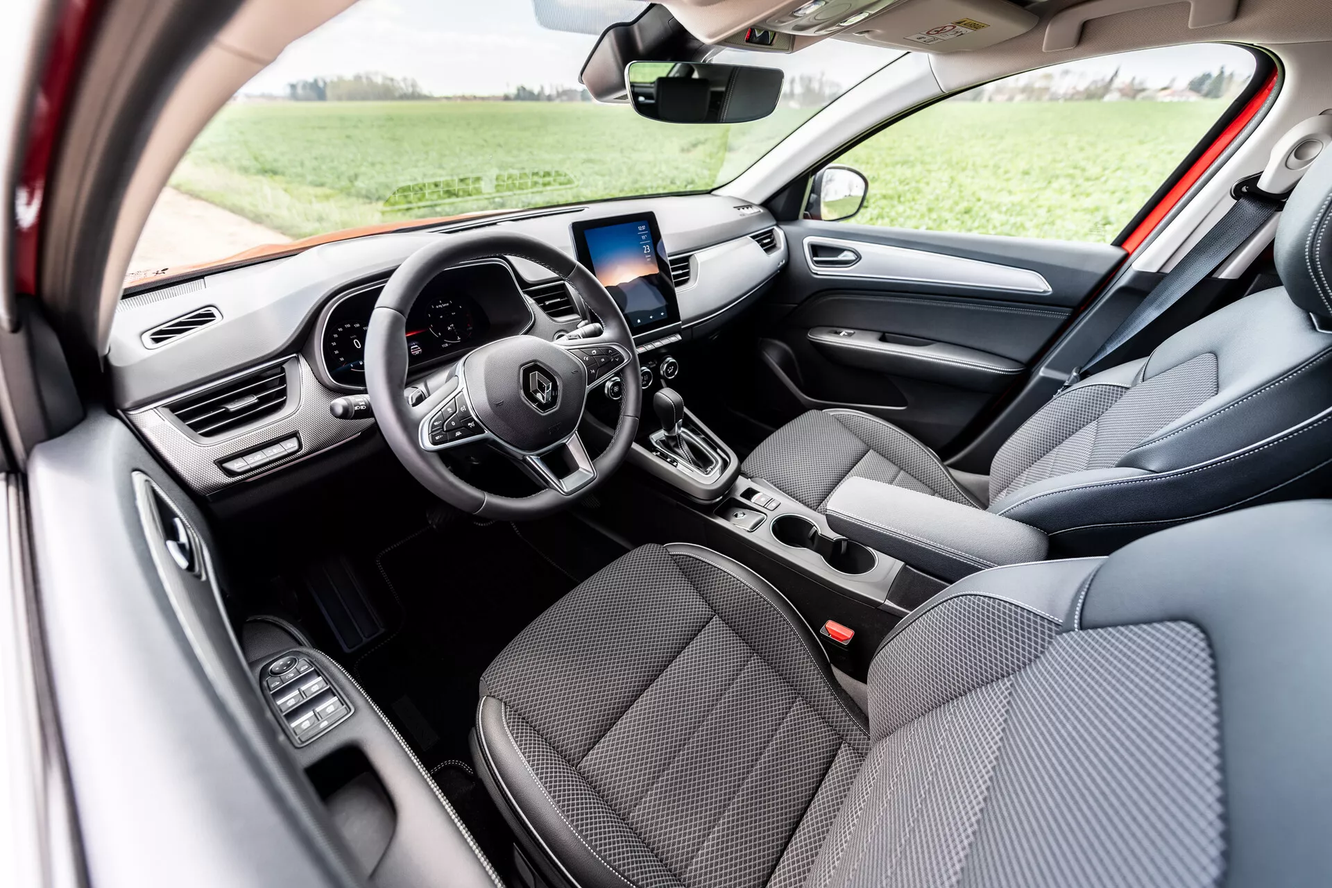 Vista interior de Renault Arkana 2021 destacando asientos delanteros en tela gris, volante multifuncional de tres radios con botones de control integrados, pantalla de tablero digital, pantalla de infoentretenimiento con navegación GPS sobre las rejillas de aire centrales, controles manuales de aire acondicionado, palanca de cambios automática y controles de ventanas eléctricas en la puerta del conductor, iluminado por luz natural diurna en un entorno exterior