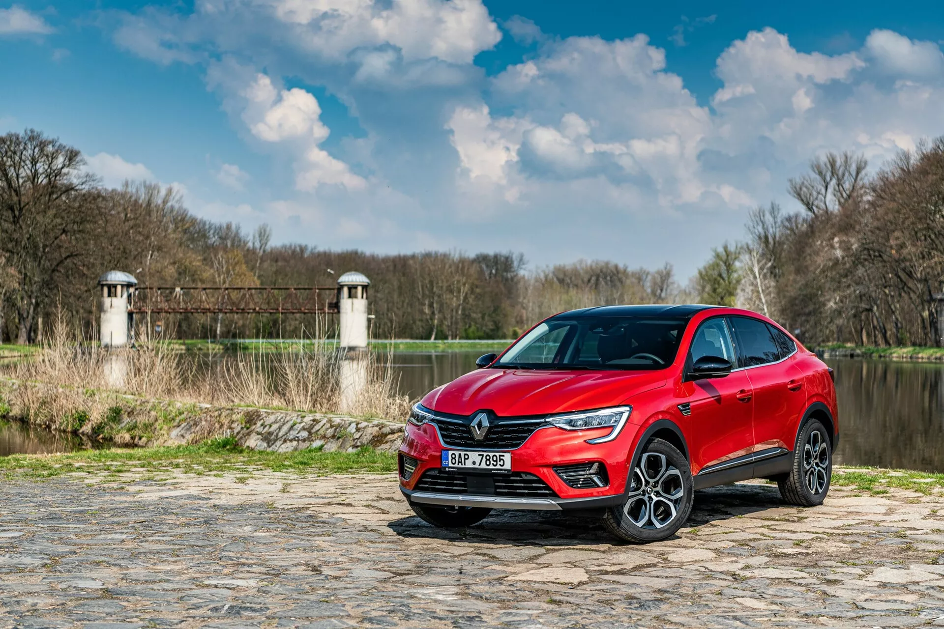 La imagen muestra un Renault Arkana 2021 estacionado en un camino de adoquines cerca de un río o lago, con vegetación y estructuras que parecen torres de una presa o puente en el fondo. El coche está capturado desde un ángulo bajo, vista frontal de tres cuartos, destacando la parrilla delantera, los faros LED y parte del perfil lateral incluyendo la puerta del conductor y el guardabarros frontal. Las características de diseño moderno, como la firma de luz nítida de los LED y las grandes ruedas de aleación, sugieren un nivel de equipamiento superior. La pintura metálica reflectante añade atractivo estético, mientras que los posibles sensores de asistencia al conductor y la tecnología incorporada en los parachoques y espejos laterales mejoran su funcionalidad.