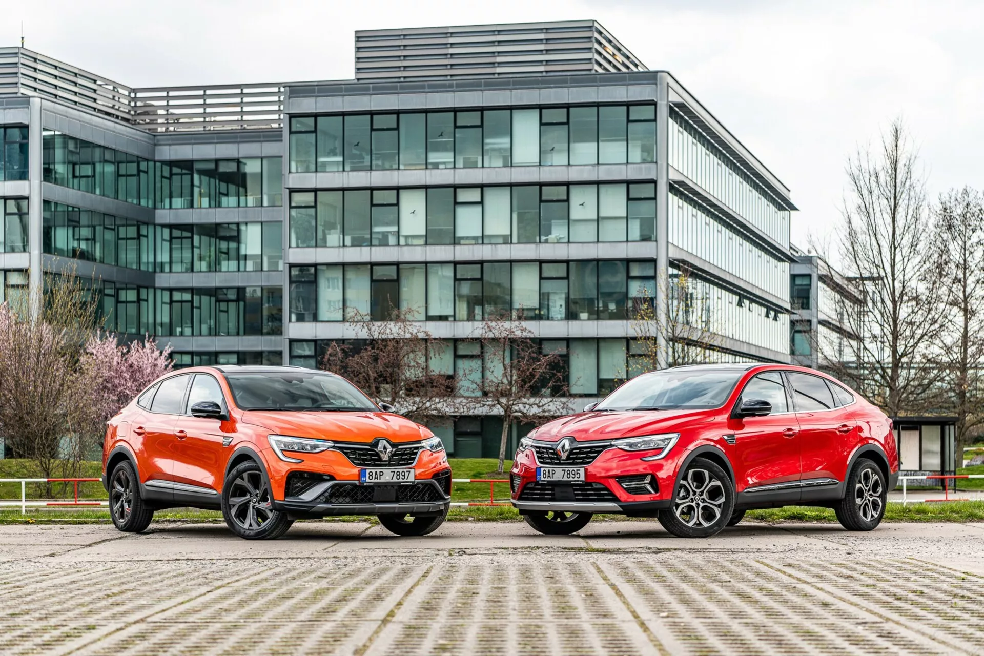 La imagen muestra dos vehículos estilo SUV estacionados lado a lado frente a un moderno edificio de oficinas con fachada de vidrio. El vehículo de la izquierda está pintado en un vibrante color naranja con un brillo metálico y presenta una parrilla frontal distintiva, faros LED nítidos, un capó esculpido y grandes ruedas de aleación multirradio con neumáticos de perfil bajo. El coche de la derecha tiene un acabado metálico rojo, con una silueta general similar, faros nítidos, una parrilla prominente y elegantes ruedas grandes de aleación. Ambos vehículos muestran líneas angulares, espejos aerodinámicos y molduras inferiores contrastantes. La foto, tomada desde un ángulo bajo cerca del nivel del suelo, subraya el entorno urbano y destaca el diseño, la presencia y las características tecnológicas avanzadas de los vehículos.