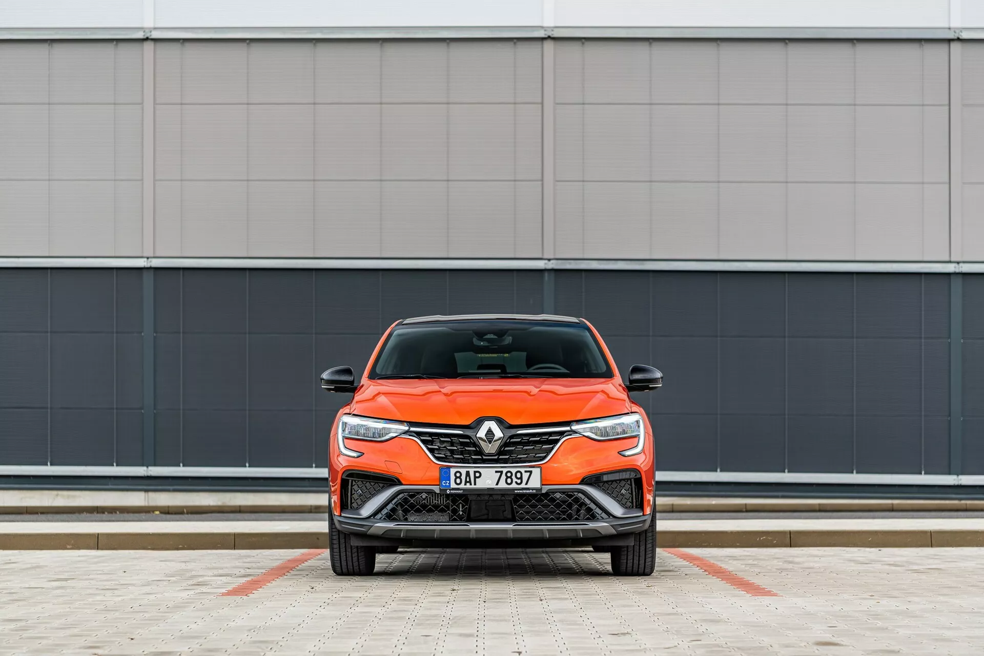 Vista frontal de un coche Renault de color naranja brillante, destacando su diseño moderno con faros elegantes y parrilla frontal, estacionado perpendicular a la cámara contra un fondo de pared gris minimalista en un área de estacionamiento lineada.
